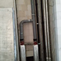 Szare rury hydrauliczne połączone kolankiem, z izolacją termiczną na dole, przymocowane do ściany z bloczków betonowych, obok inne rury w pionie.