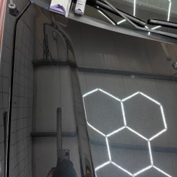 GregorDetailing - Błyszcząca, czarna karoseria samochodu po nałożeniu powłoki ceramicznej, widoczne opakowanie produktu UltraCoat Carbon i odbicie osoby robiącej zdjęcie w lakierze, tło stanowią heksagonalne lampy...