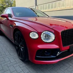 Czerwony Bentley Flying Spur z czarnymi felgami i detalami, zaparkowany na szarym bruku, z widoczną częścią budynku w tle, prezentujący luksusowy design.