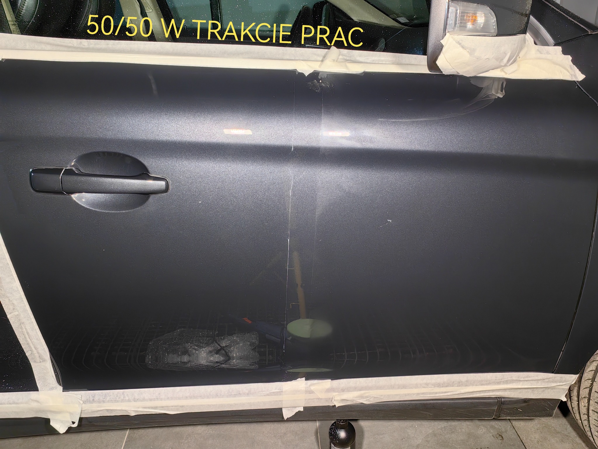 Część karoserii samochodu w trakcie polerowania, widoczna różnica w połysku między obszarem po lewej i prawej stronie, zabezpieczona taśmą malarską, napis '50/50 W TRAKCIE PRAC' na szybie.