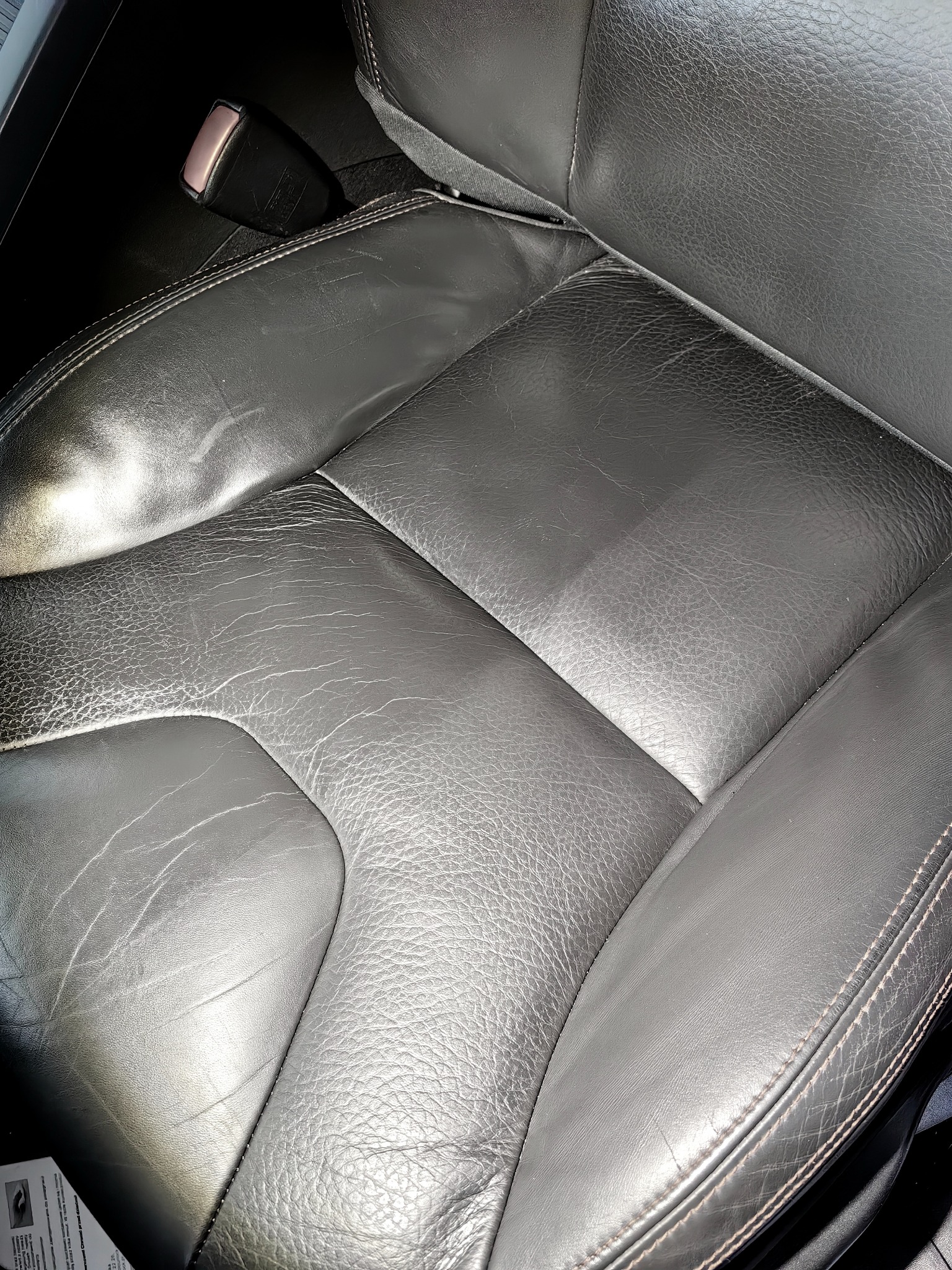 Fragment czarnego skórzanego fotela samochodowego z widocznymi śladami użytkowania i zapięciem pasów bezpieczeństwa.