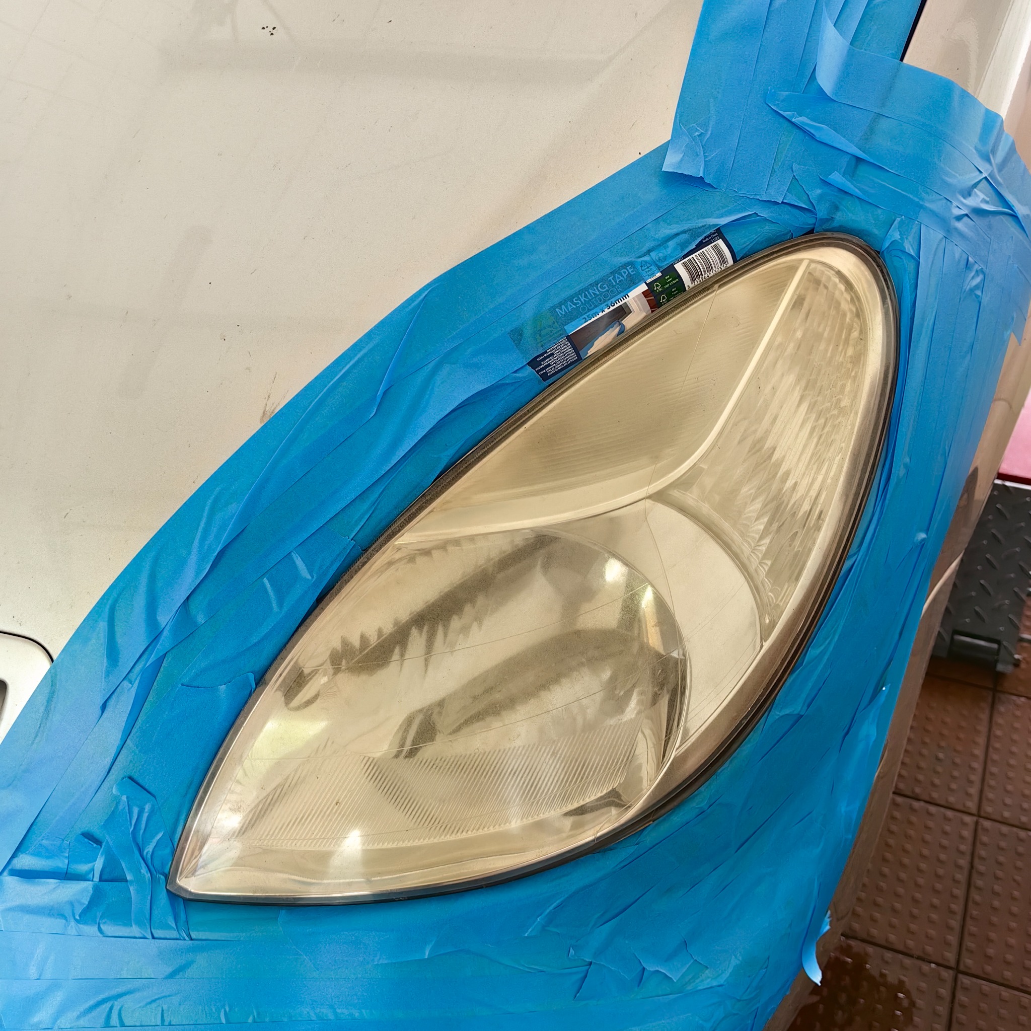 Przygotowanie reflektora samochodowego do polerowania, zabezpieczonego niebieską taśmą maskującą, widoczny fragment opakowania taśmy.