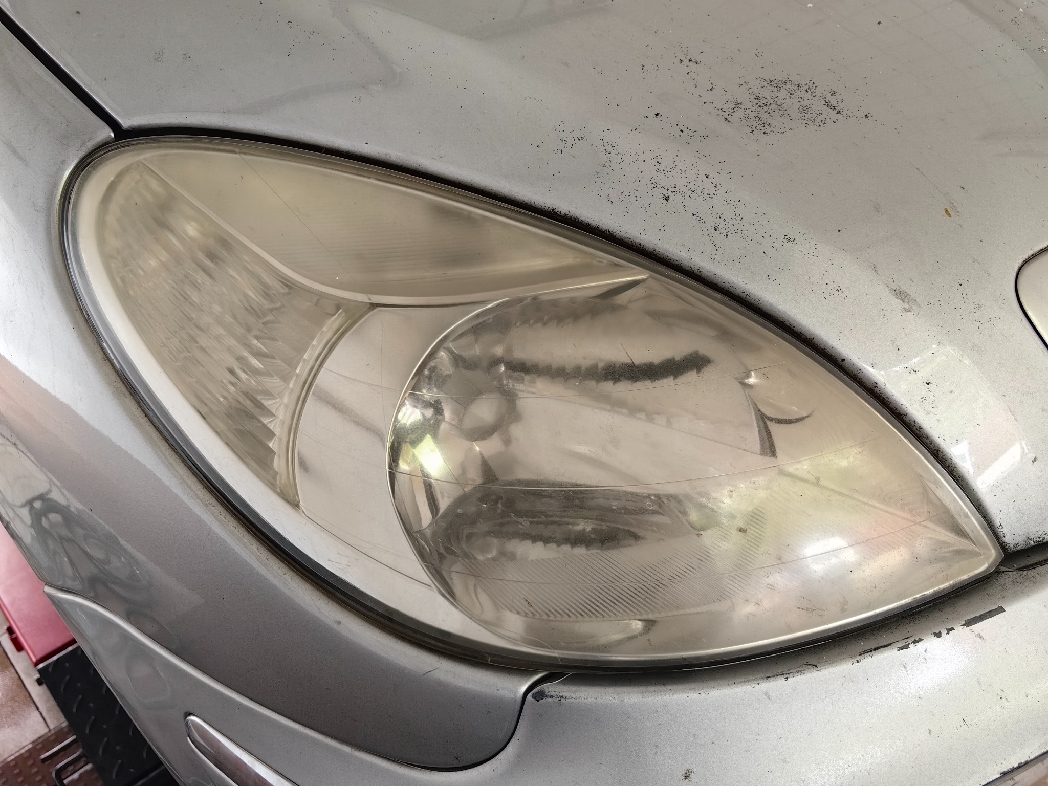 Przednia lampa samochodu w kolorze srebrnym z widocznymi śladami zabrudzeń i zmatowienia na kloszu, wymagająca polerowania lub regeneracji.