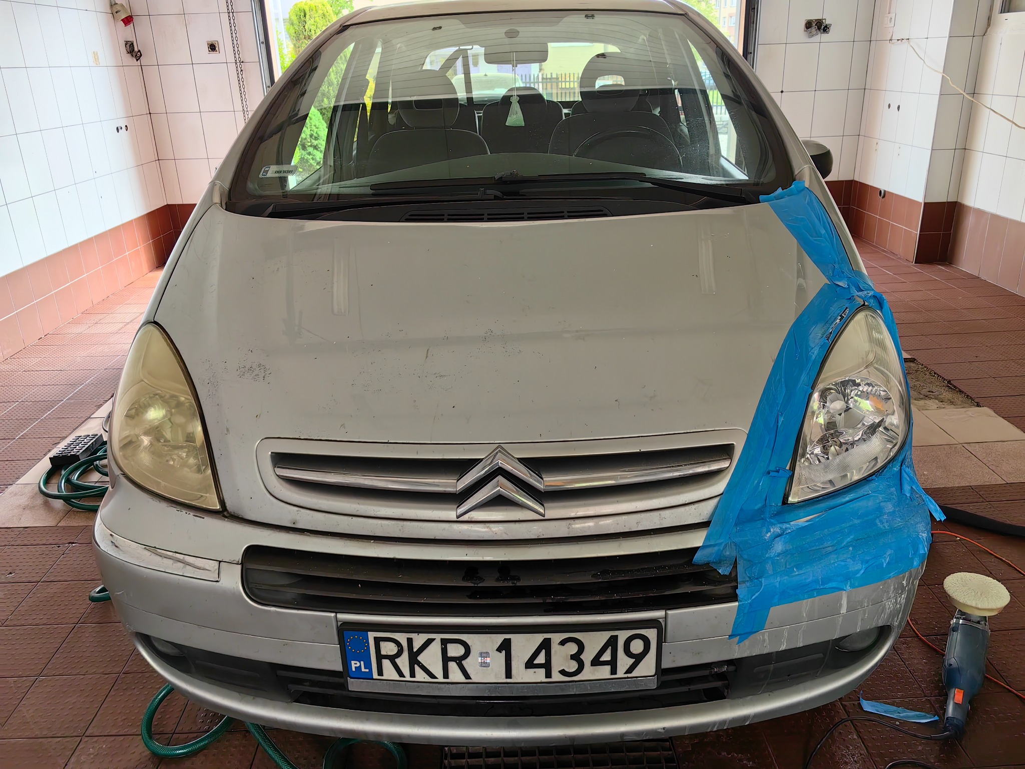 Srebrny Citroen Picasso z zaklejoną taśmą jedną lampą przednią, przygotowany do polerowania, z widoczną polerką obok.
