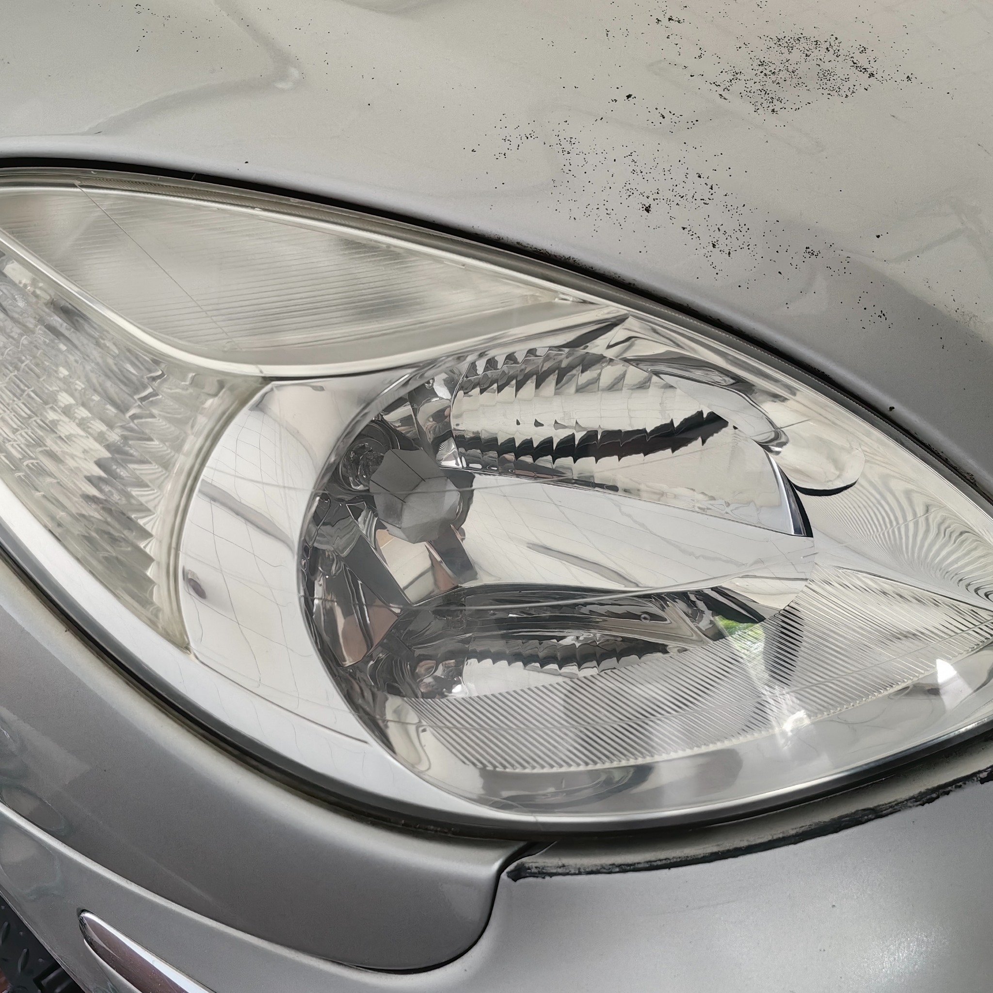 Szczegółowe ujęcie przedniego reflektora srebrnego samochodu z widocznymi śladami zabrudzeń na karoserii powyżej lampy.