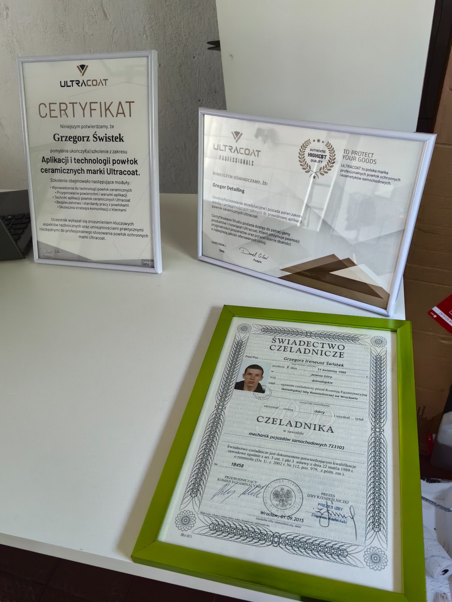 Trzy certyfikaty w ramkach na białym stole, potwierdzające kwalifikacje i szkolenia z zakresu aplikacji powłok ceramicznych marki Ultracoat dla Grzegorza Świstka i Gregor Detailing, w tym...