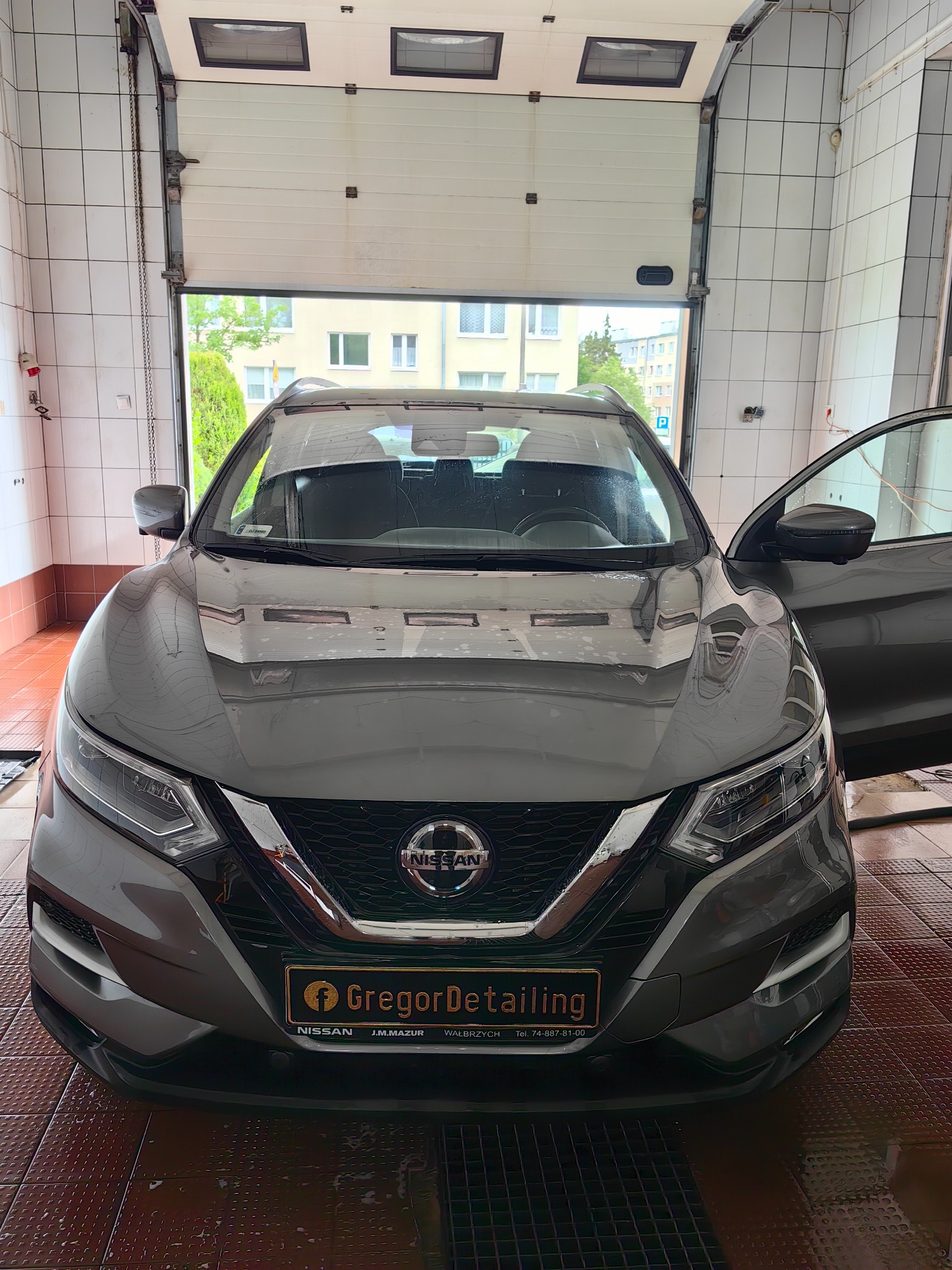 Szary Nissan Qashqai w myjni samochodowej z otwartymi drzwiami, widoczna tabliczka z logo firmy detailingowej na przednim zderzaku, ceramiczne płytki na ścianach i podłodze.