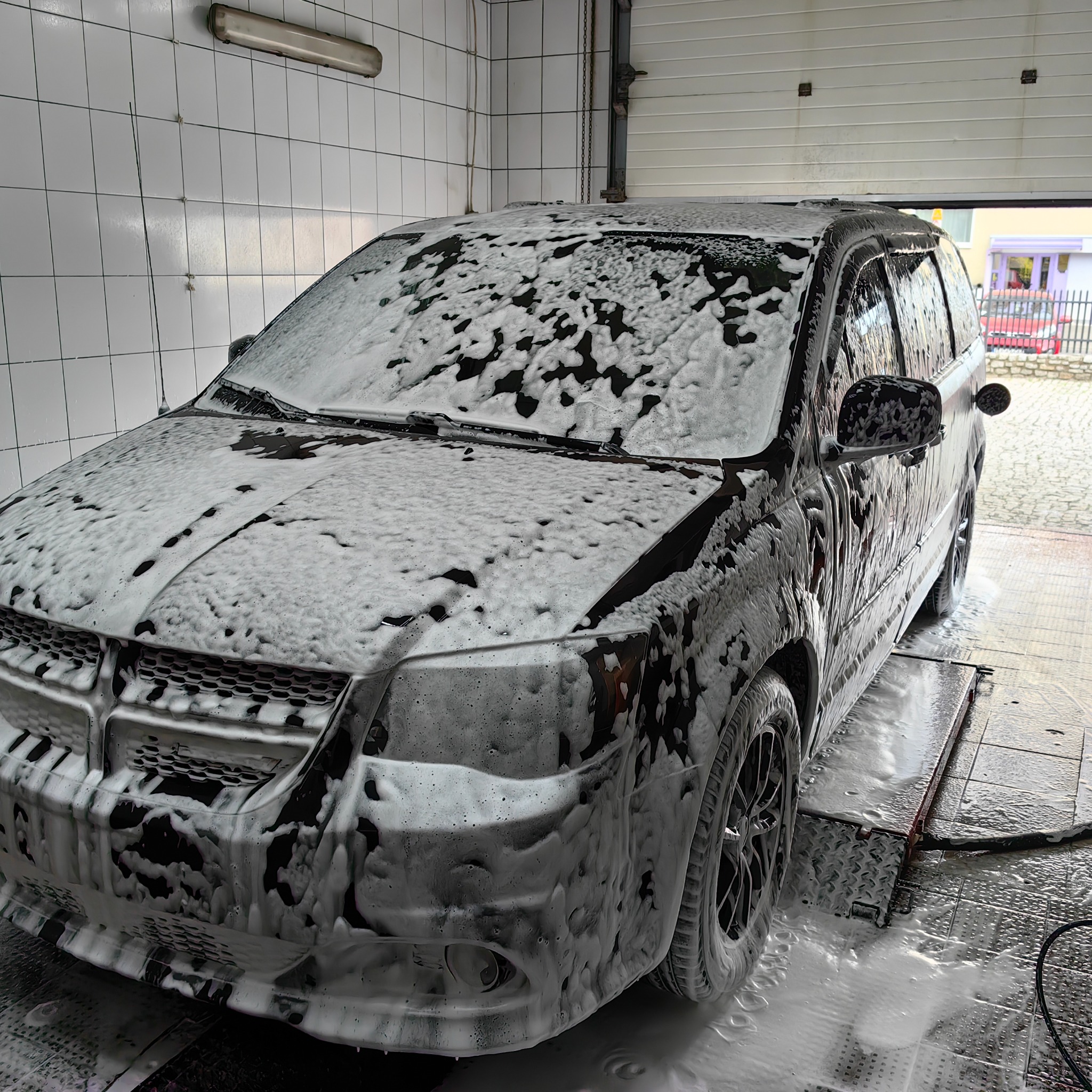 Czarny samochód typu minivan pokryty pianą w myjni, widok z przodu, otwarte drzwi garażowe w tle.