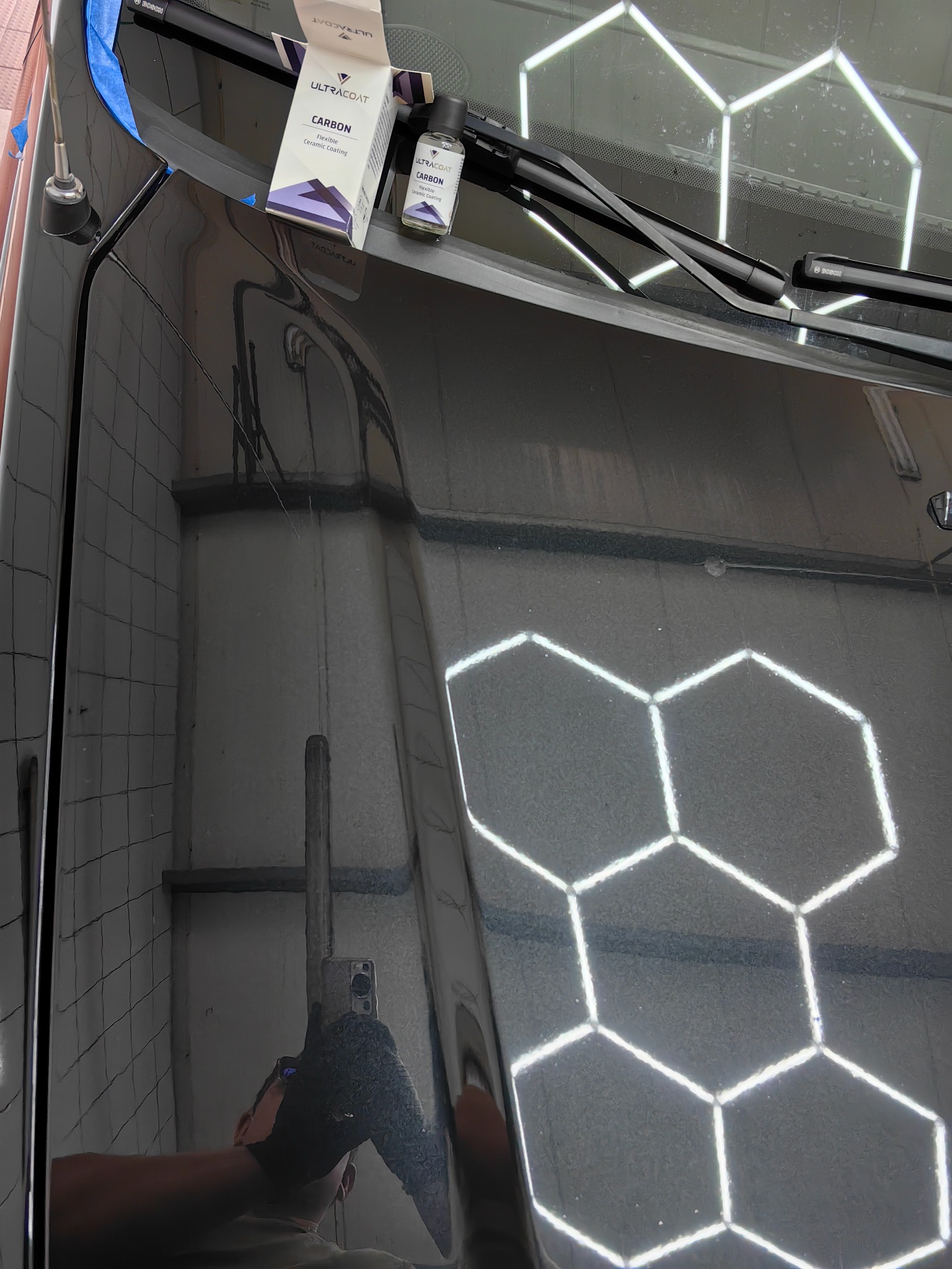 Błyszcząca, czarna karoseria samochodu po nałożeniu powłoki ceramicznej, widoczne opakowanie produktu UltraCoat Carbon i odbicie osoby robiącej zdjęcie w lakierze, tło stanowią heksagonalne lampy...