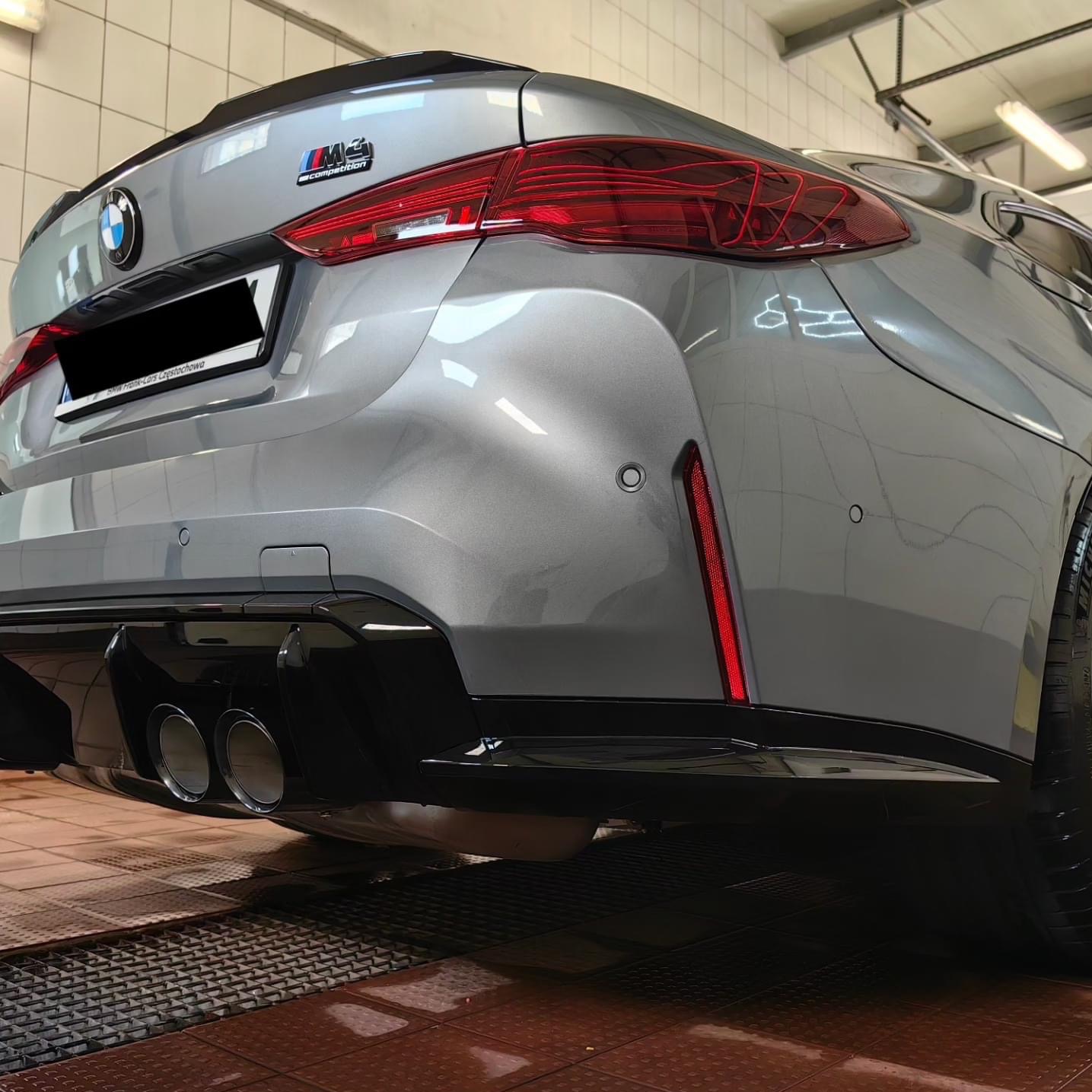 Tył szarego samochodu BMW M3 Competition z czarnym dyfuzorem i podwójnymi końcówkami wydechu, widoczny w myjni z kratką podłogową.