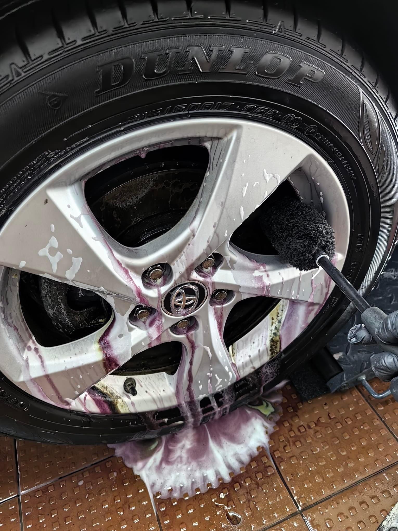 Czyszczenie aluminiowej felgi samochodowej szczotką, spływający fioletowy płyn z pianą na brązową kostkę brukową.