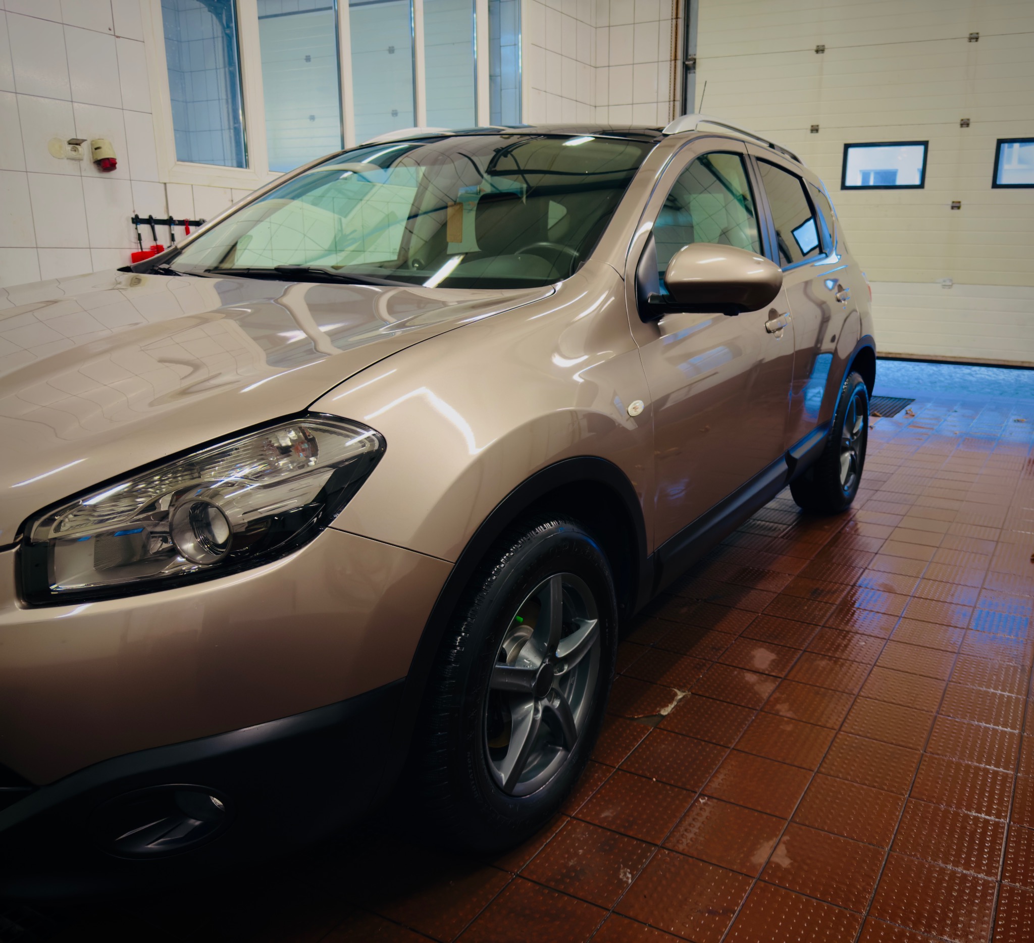 Czysty, beżowy Nissan Qashqai po myciu w myjni. Widoczne mokre, brązowe płytki podłogowe i biała brama garażowa w tle. Lśniące reflektory i opony.