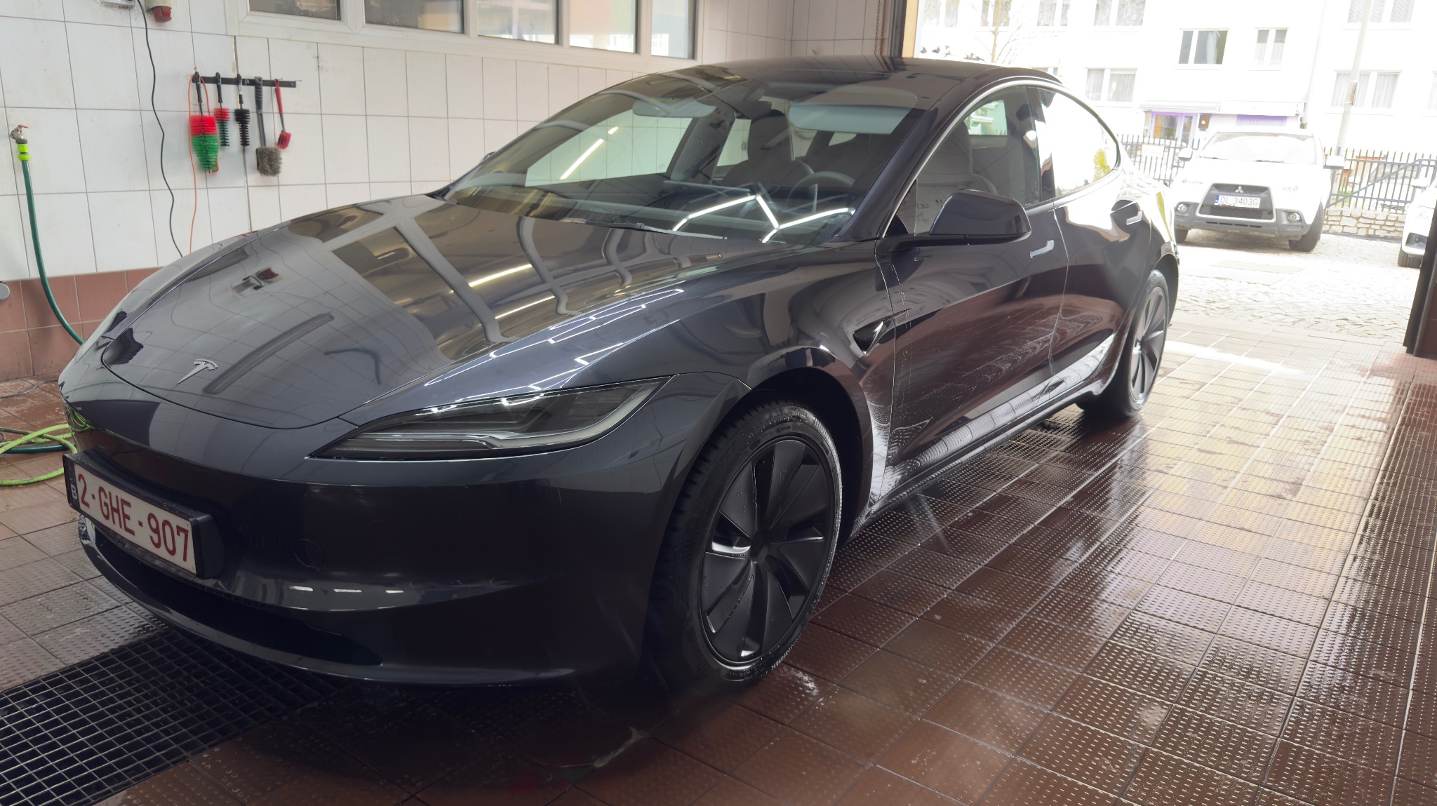 Błyszcząca, ciemnoszara Tesla Model 3 po myciu w myjni, z widocznym numerem rejestracyjnym i mokrą podłogą z płytek. W tle widoczne narzędzia do mycia i inny samochód.
