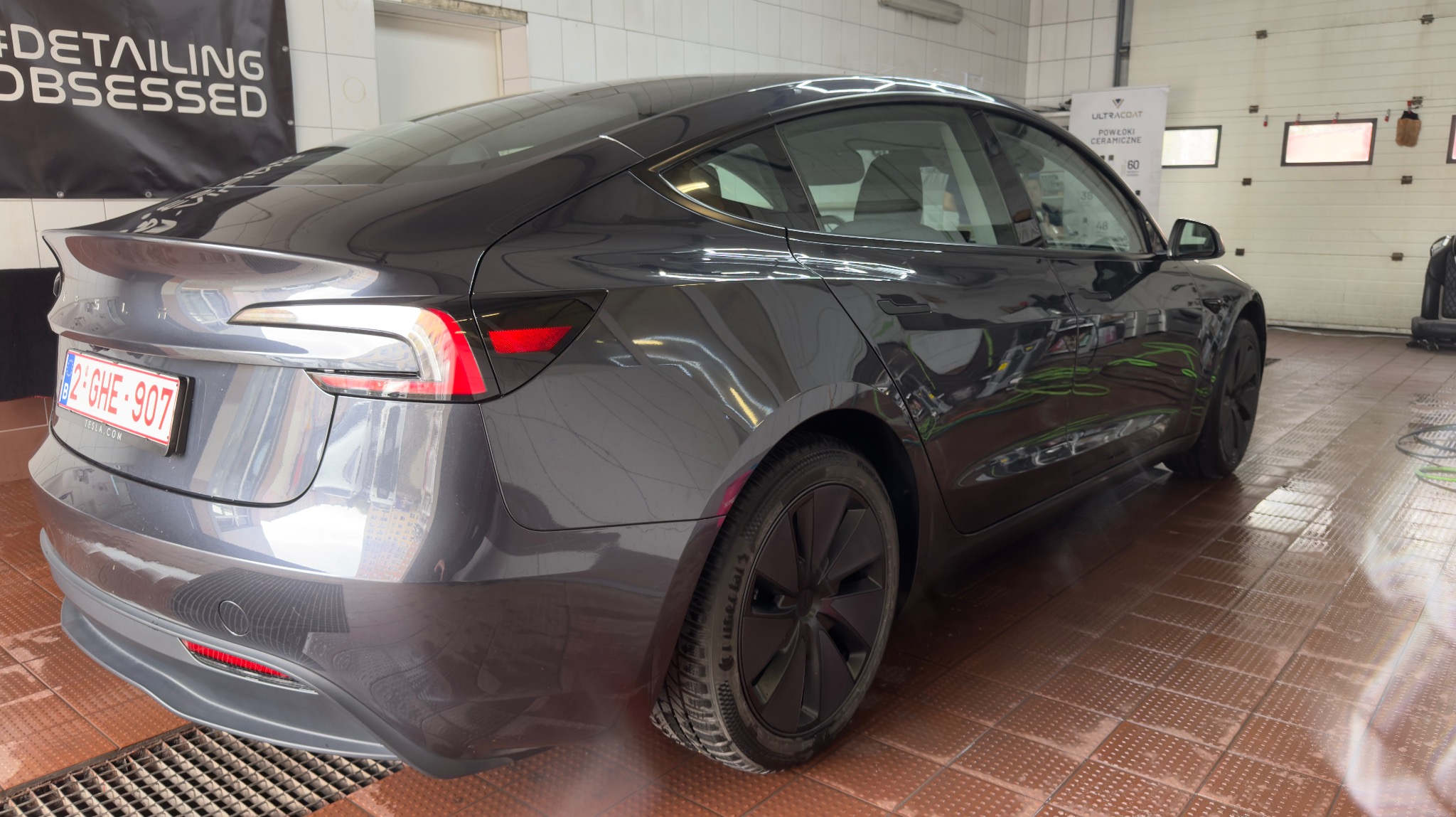 Błyszcząca, szara Tesla Model 3 po detailingu, widziana z tyłu pod kątem. Widoczny napis #DETAILINGOBSESSED oraz logo UltraCoat na ścianie myjni.
