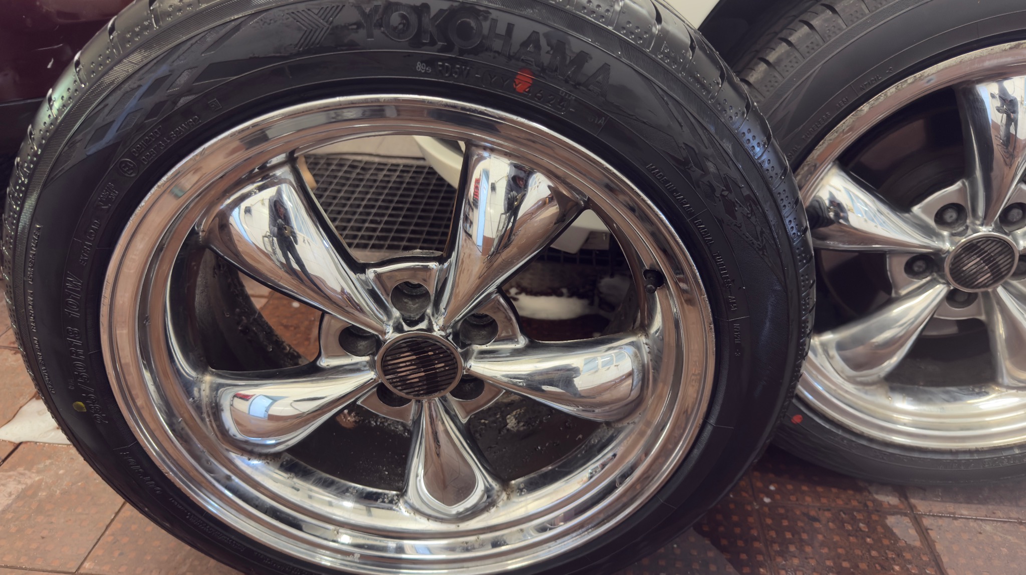 Dwa chromowane felgi Yokohama z oponami, widoczne detale bieżnika i wzór felgi. Odbicia w polerowanej powierzchni. Układ kół na brązowej, kwadratowej powierzchni.