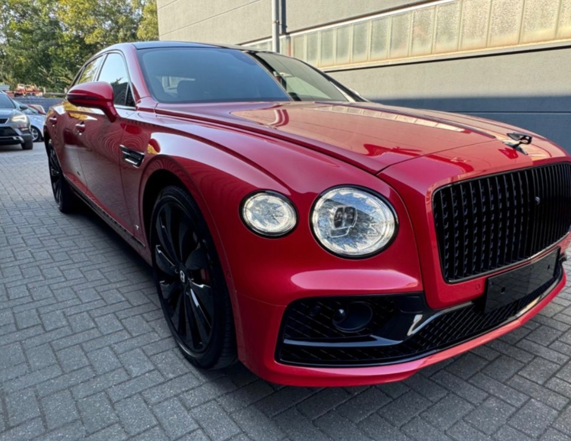 Czerwony Bentley Flying Spur z czarnymi felgami i detalami, zaparkowany na szarym bruku, z widoczną częścią budynku w tle, prezentujący luksusowy design.
