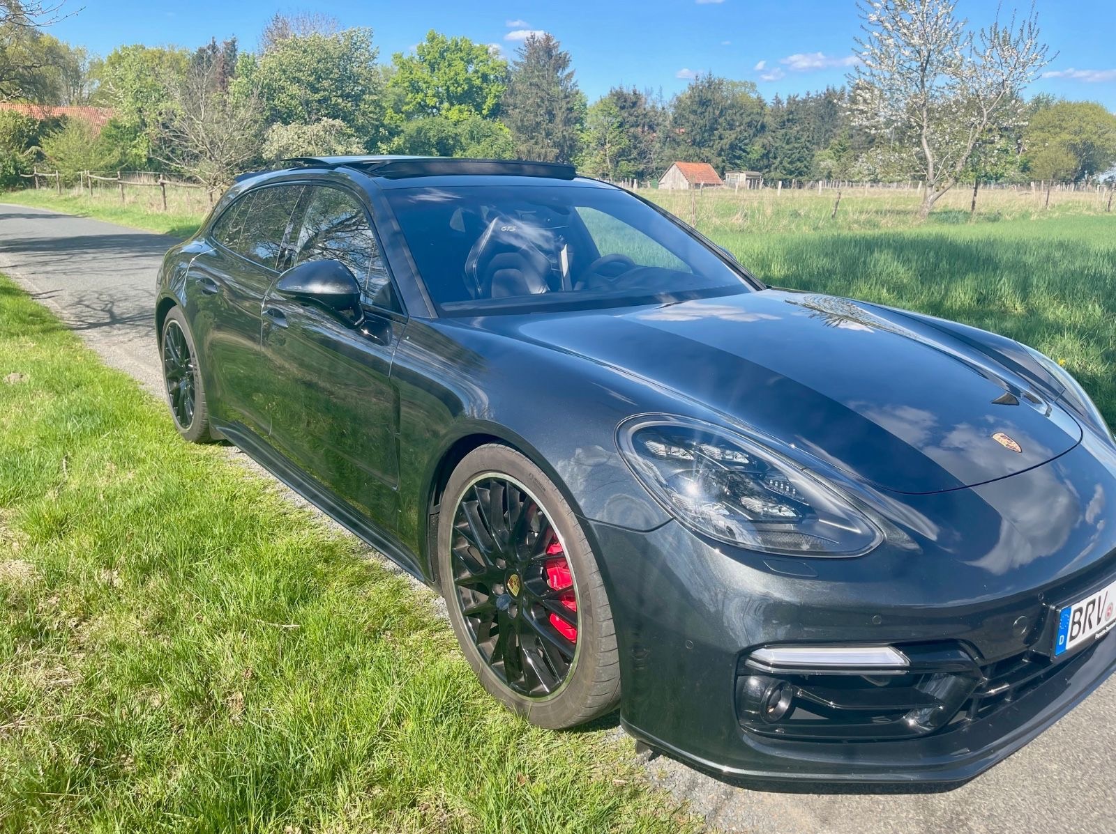 Czarny Porsche Panamera Sport Turismo na trawie, z widocznymi czerwonymi zaciskami hamulcowymi i czarnymi felgami. Słoneczny dzień, drzewa w tle.