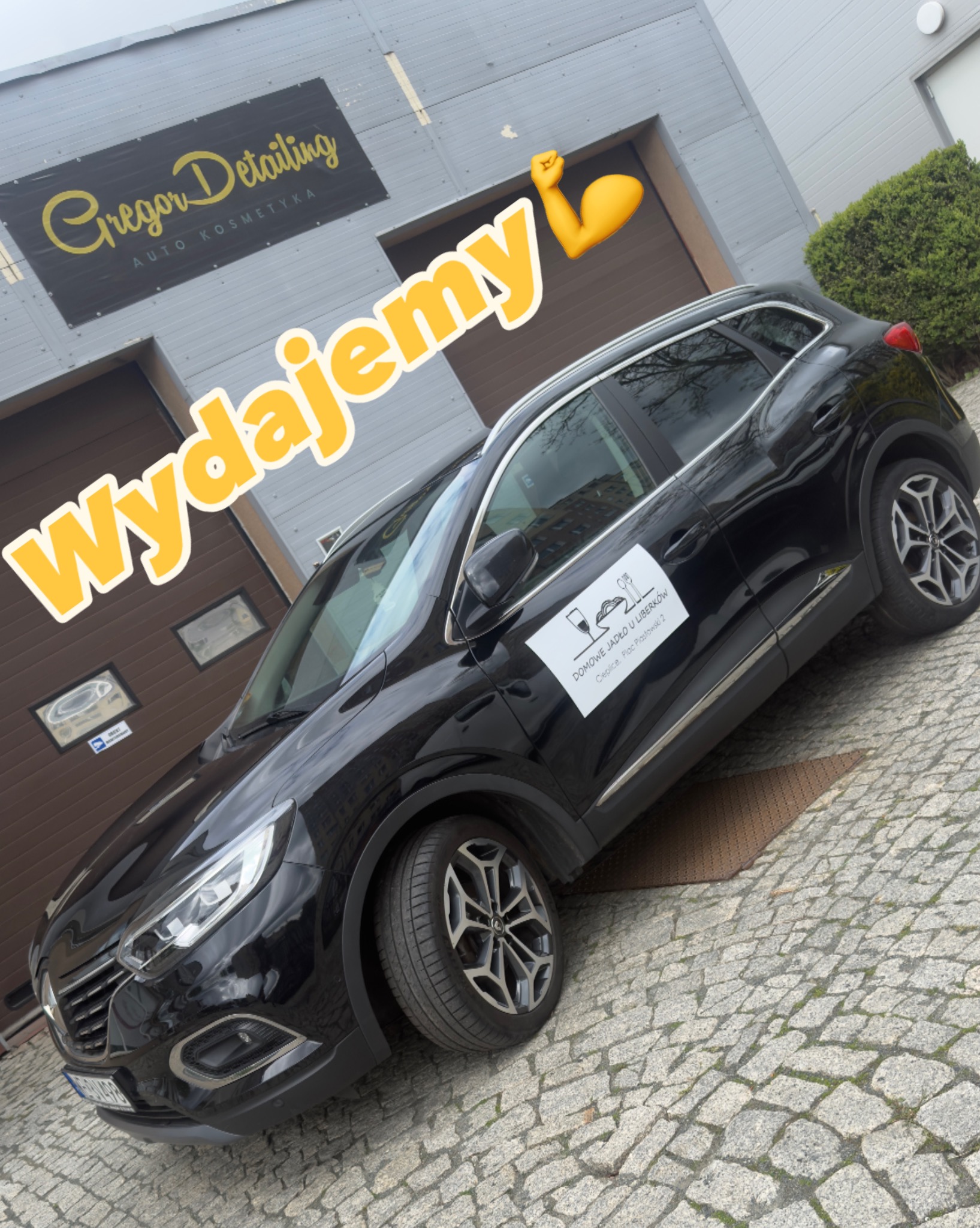 Czarny SUV po detailingu przed budynkiem z szyldem 'Gregor Detailing', z naklejką restauracji na drzwiach. Ujęcie z dołu, z żółtym napisem 'Wydajemy'.