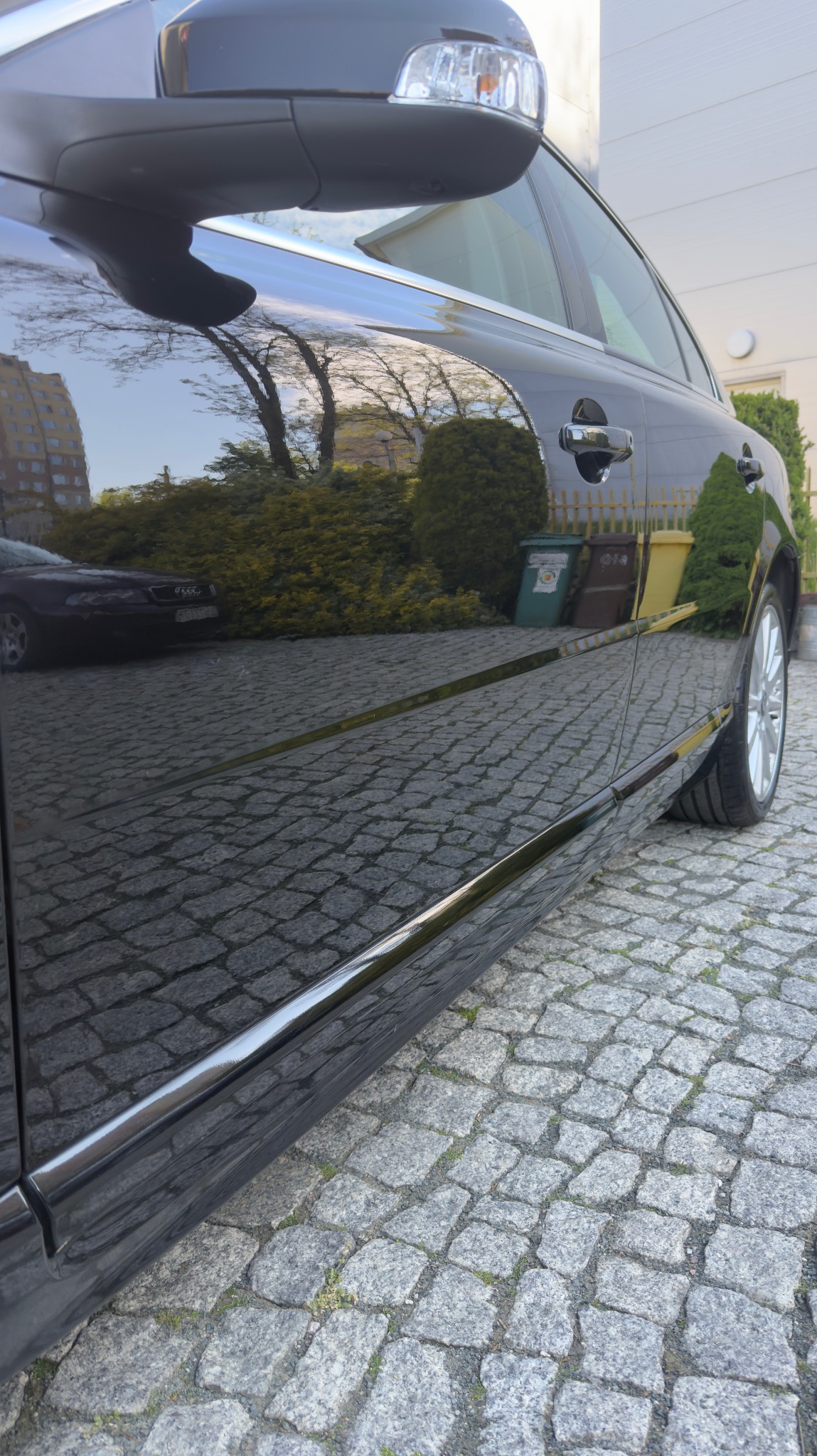 Błyszczący, czarny samochód zaparkowany na brukowanej nawierzchni. W lakierze odbija się budynek i zieleń, co podkreśla efekt lustrzanego połysku po umyciu.