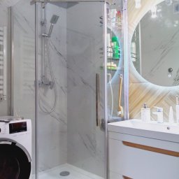 Usługi remontowo budowlane - Nowoczesna kabina prysznicowa z deszczownicą, umieszczona w łazience wykończonej marmurowymi płytkami, obok pralki i umywalki z szafką z drewnianymi akcentami oraz okrągłym lustrem z podświetleniem...