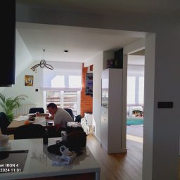 Usługi remontowo budowlane - Wnętrze domu w trakcie remontu: mężczyzna przy stole omawia plany, widoczna kuchnia z wyspą, jadalnia i fragment salonu z ceglaną ścianą, elementy wyposażenia zabezpieczone folią.