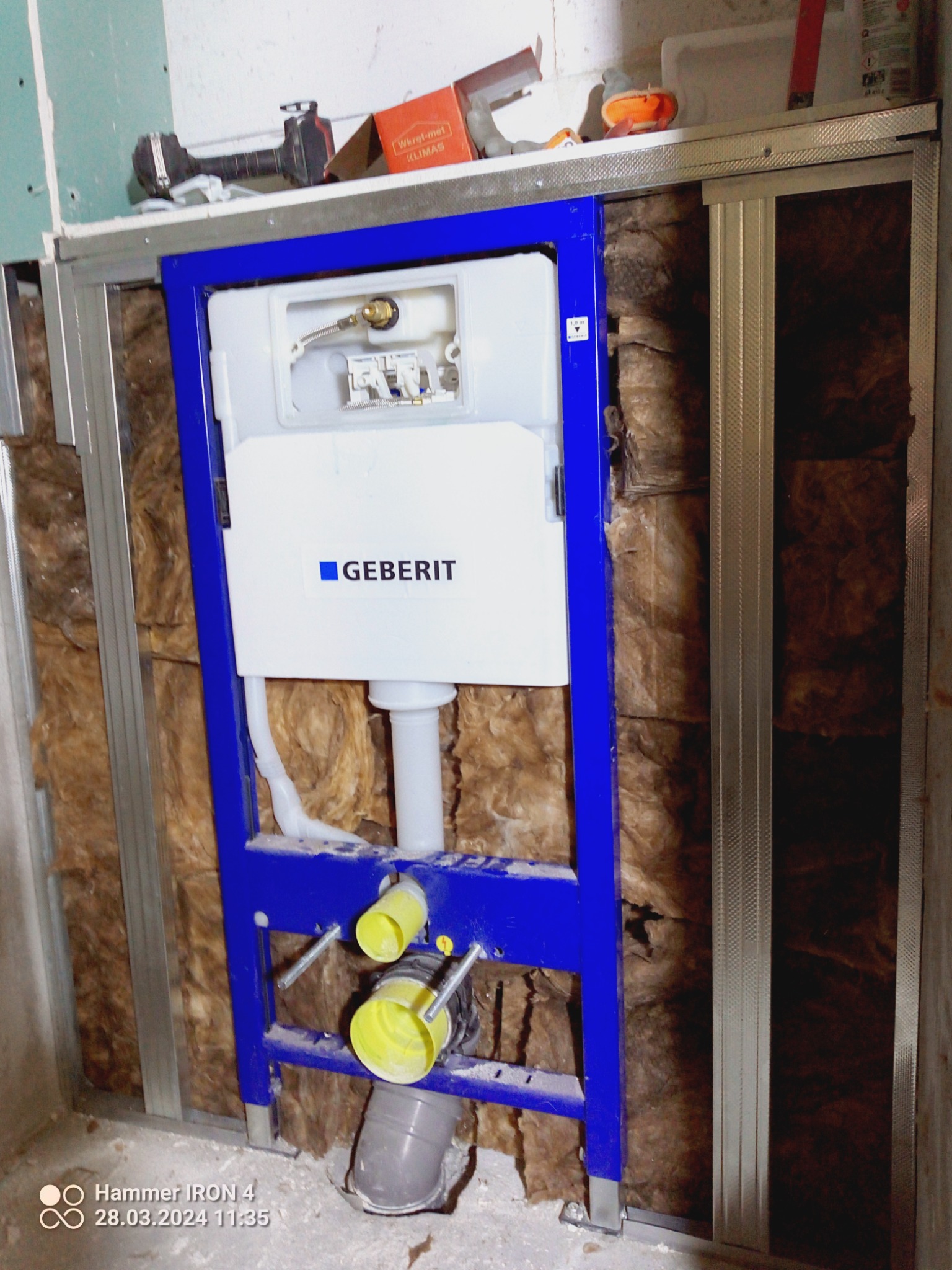 Stelaż podtynkowy Geberit zamontowany w łazience, widoczne rury odpływowe, izolacja z wełny mineralnej i metalowa konstrukcja szkieletowa ściany działowej, w tle narzędzia.