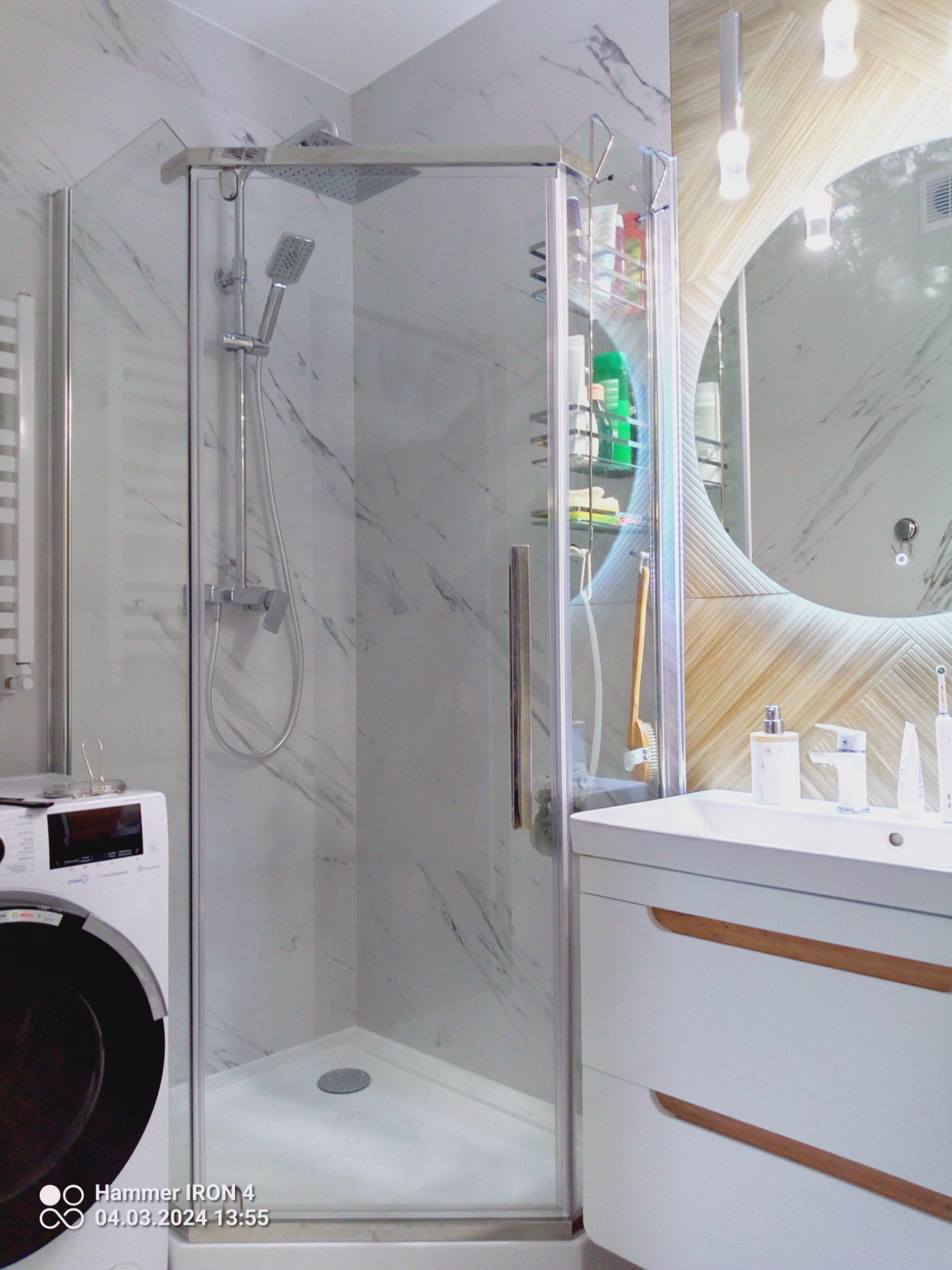 Nowoczesna kabina prysznicowa z deszczownicą, umieszczona w łazience wykończonej marmurowymi płytkami, obok pralki i umywalki z szafką z drewnianymi akcentami oraz okrągłym lustrem z podświetleniem...