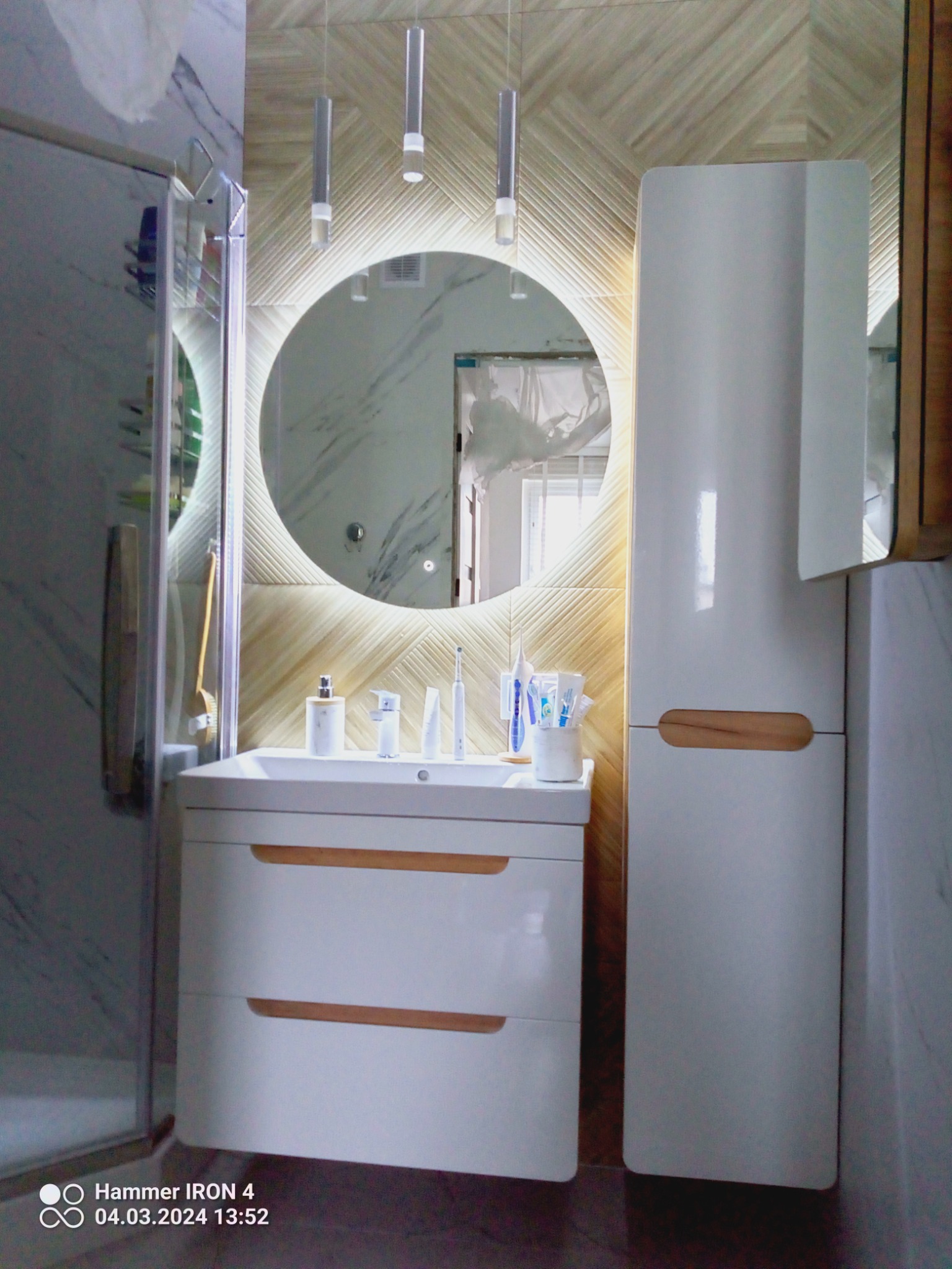 Nowoczesna łazienka z okrągłym lustrem podświetlanym LED, białą szafką pod umywalką z drewnianymi akcentami i wysoką szafką, widoczne płytki imitujące drewno na ścianie i marmur na sąsiedniej...