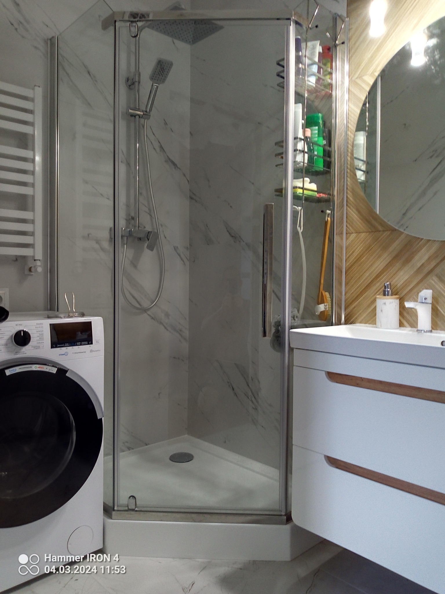 Nowoczesna łazienka z kabiną prysznicową narożną z przezroczystego szkła, białą umywalką z drewnianymi akcentami, pralką oraz grzejnikiem drabinkowym na ścianie wyłożonej płytkami imitującymi marmur.