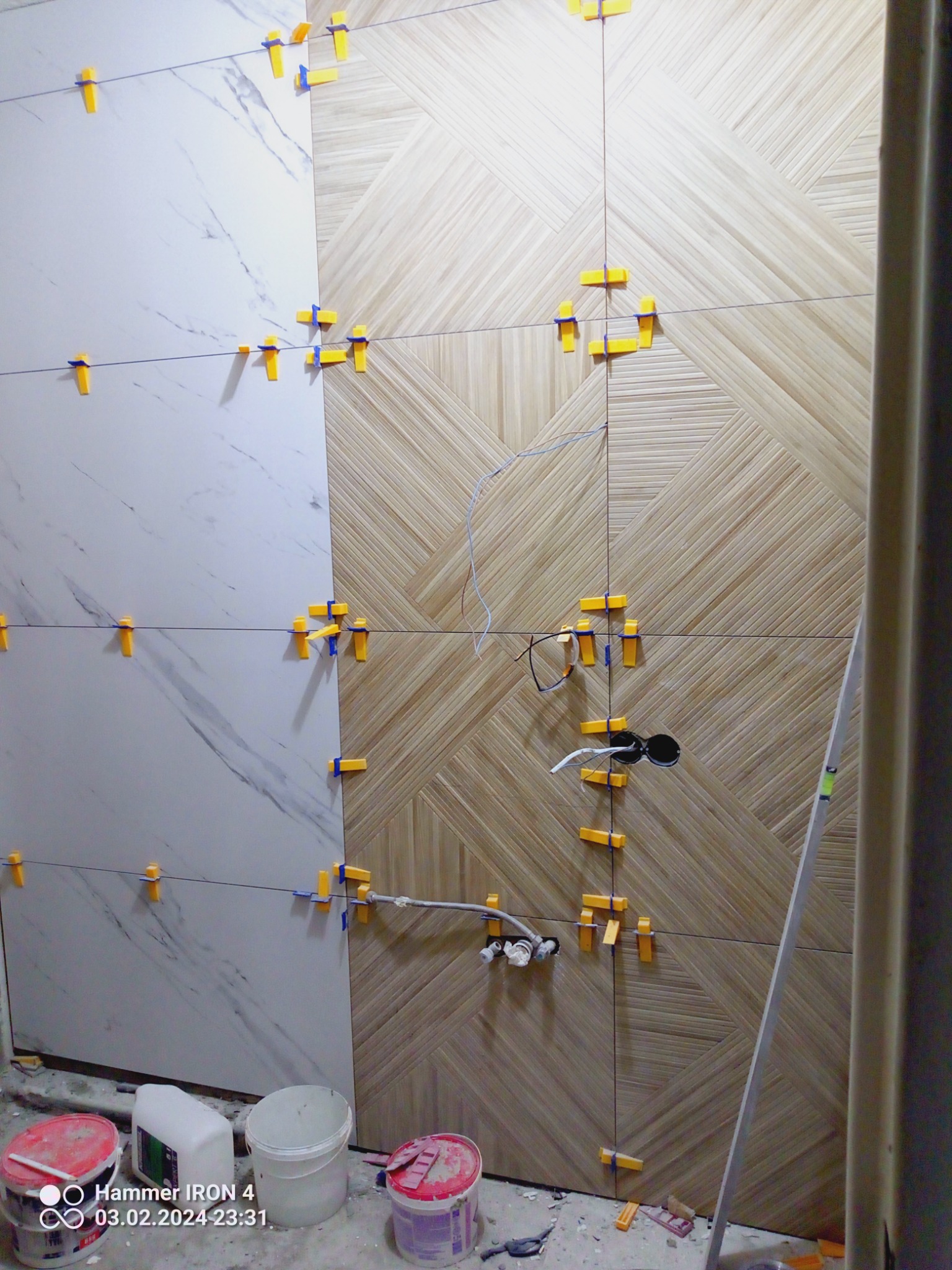 Układanie płytek na ścianie, widoczne płytki imitujące marmur i drewno z użyciem poziomujących klipsów, puszka elektryczna i rury hydrauliczne w ścianie, na podłodze wiadra i narzędzia.