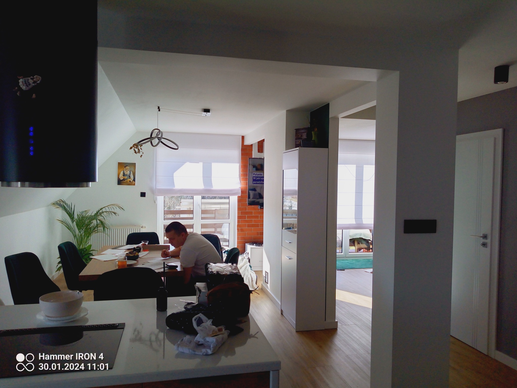 Wnętrze domu w trakcie remontu: mężczyzna przy stole omawia plany, widoczna kuchnia z wyspą, jadalnia i fragment salonu z ceglaną ścianą, elementy wyposażenia zabezpieczone folią.