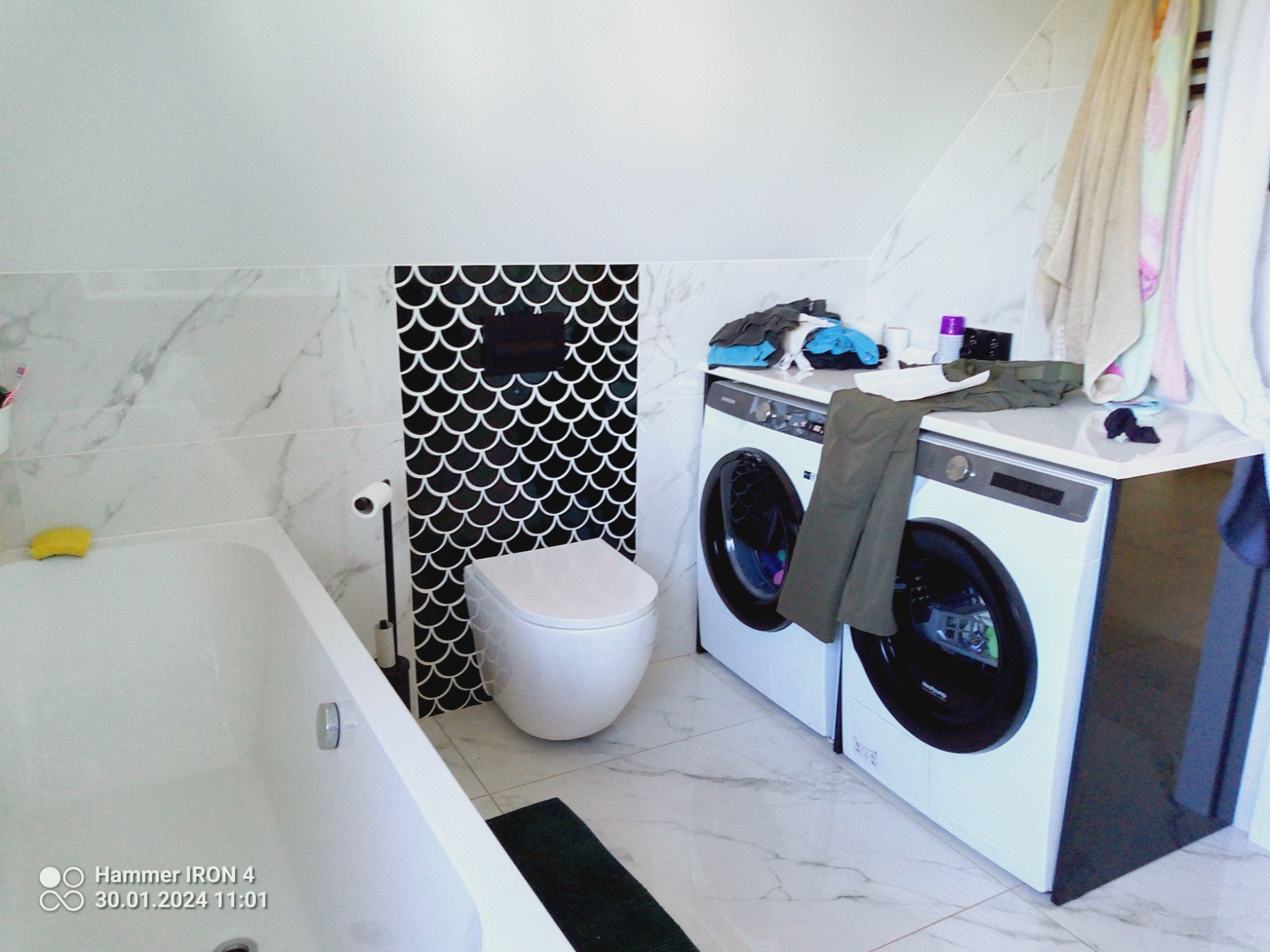 Nowoczesna łazienka na poddaszu z białą wanną, toaletą podwieszaną z czarną mozaiką w kształcie rybiej łuski, pralką i suszarką obok siebie oraz marmurowymi płytkami na podłodze i ścianach.