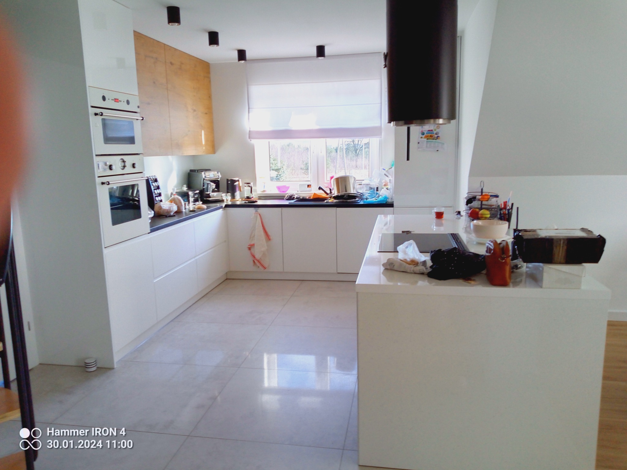 Jasna kuchnia z białymi meblami, drewnianymi akcentami i oknem z roletą rzymską, widok na zewnątrz