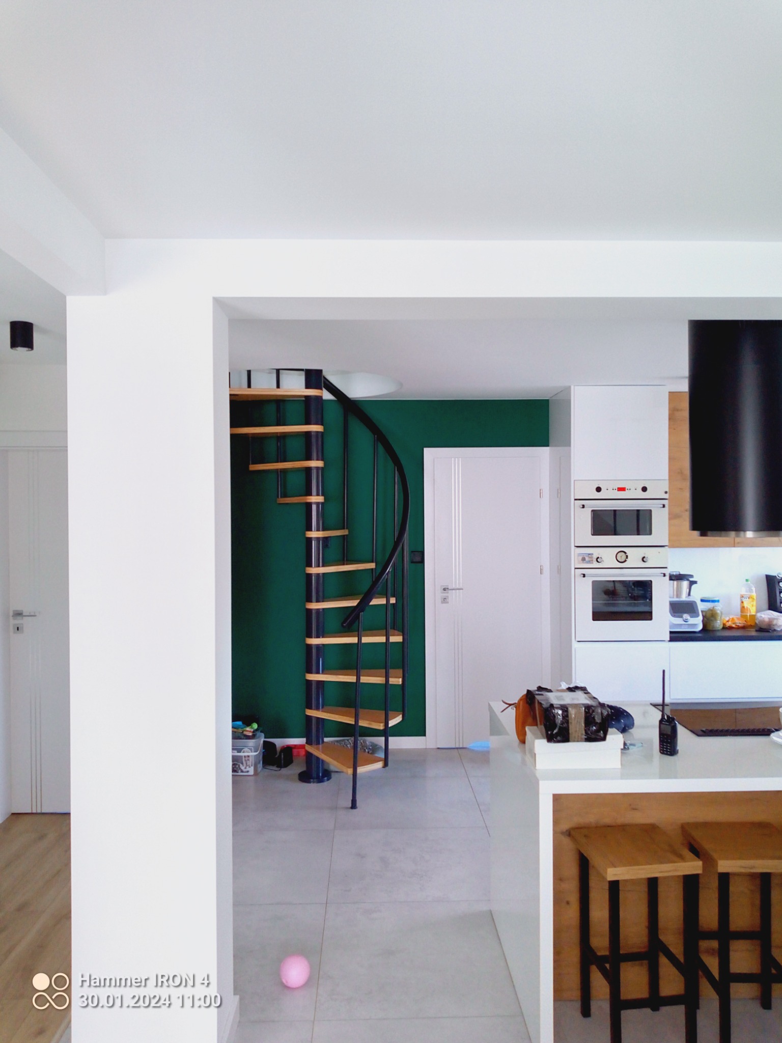 Wnętrze mieszkania z spiralnymi schodami prowadzącymi na wyższą kondygnację, widok z salonu na kuchnię z wyspą i hokerami, ściana pomalowana na ciemny zielony kolor.