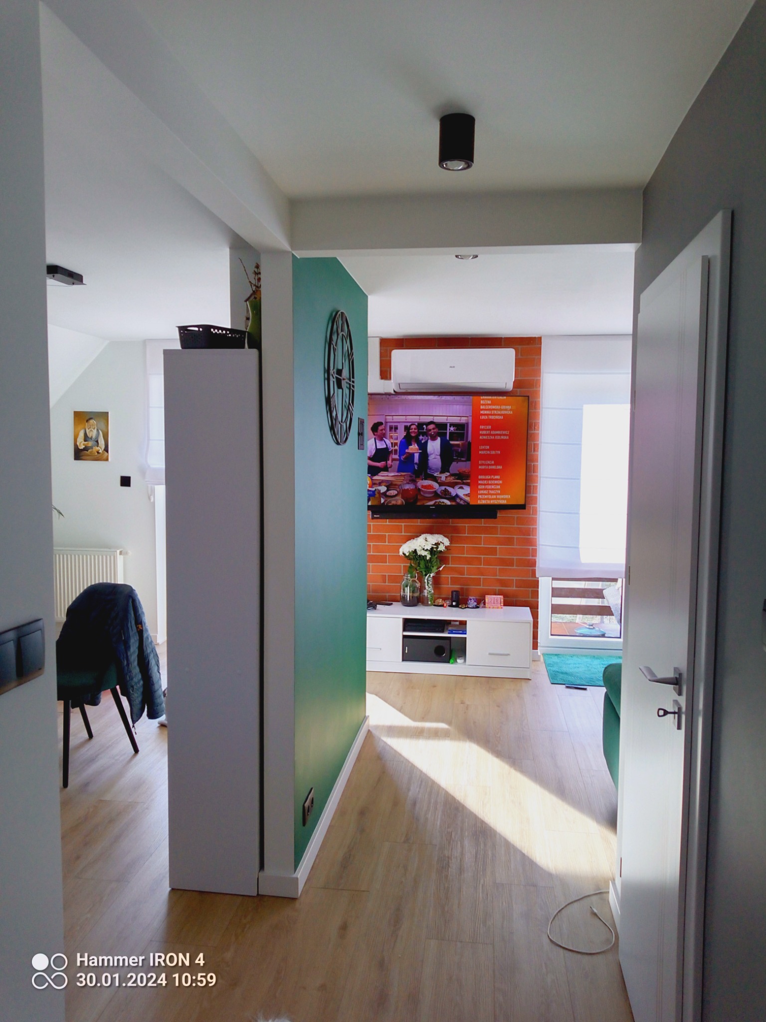Wnętrze po odświeżeniu w Bełchatowie: widok z korytarza na salon z ceglaną ścianą, telewizorem, białymi meblami i drewnianą podłogą, z akcentami turkusowej ściany i dekoracyjnym zegarem.