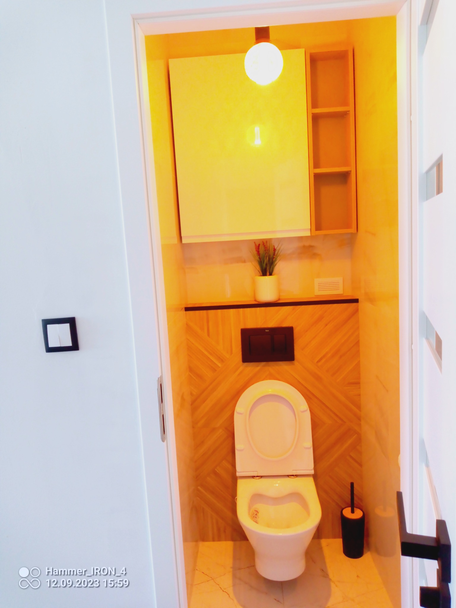 Jasna łazienka z toaletą, drewnianą mozaiką na ścianie, szafką nad toaletą i marmurową podłogą, oświetlona ciepłym światłem