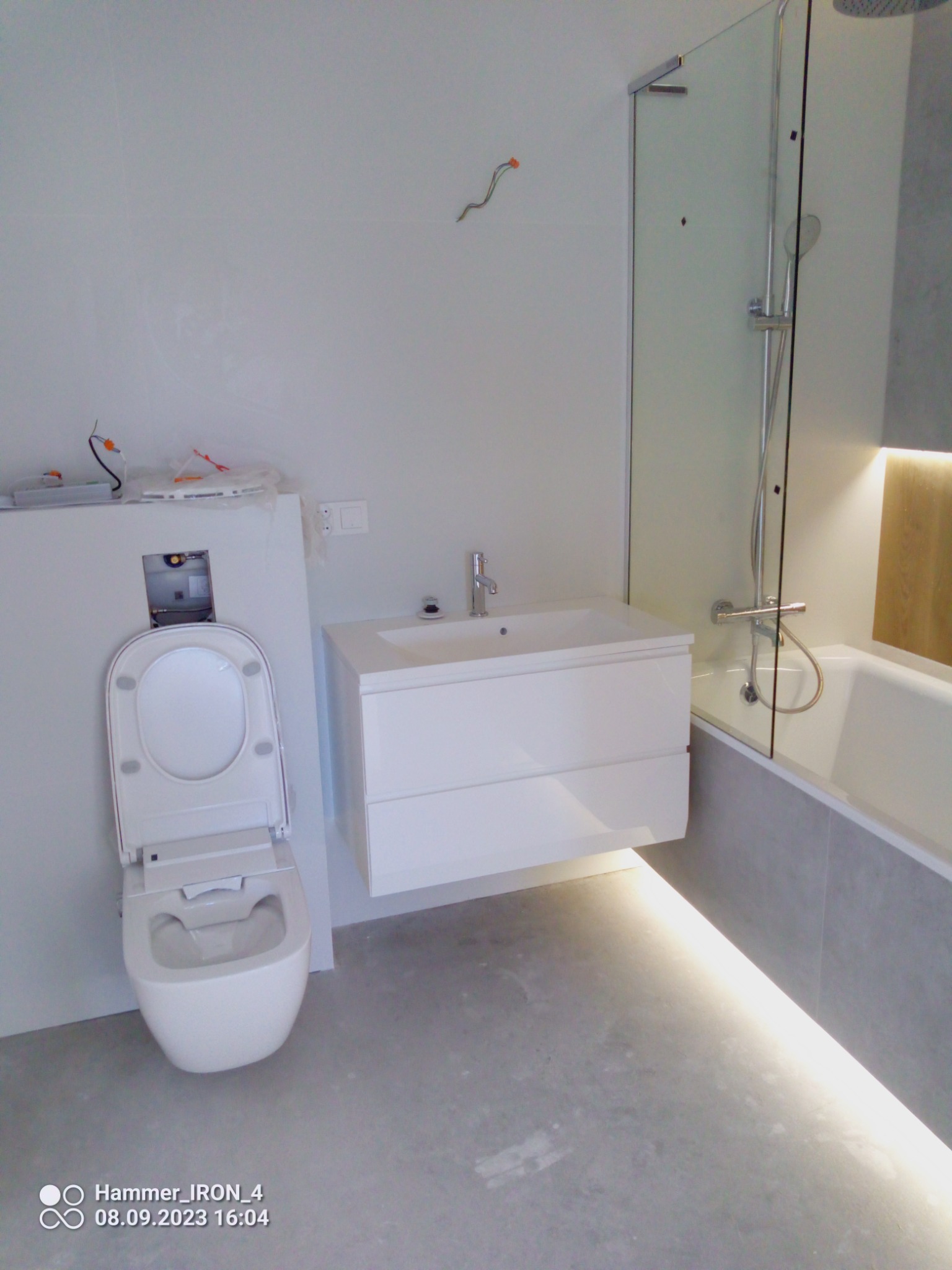 Nowoczesna łazienka w trakcie wykańczania: podwieszana toaleta z otwartą deską, umywalka z szafką i podświetleniem LED pod spodem, szklana kabina prysznicowa z chromowaną armaturą, widoczne...