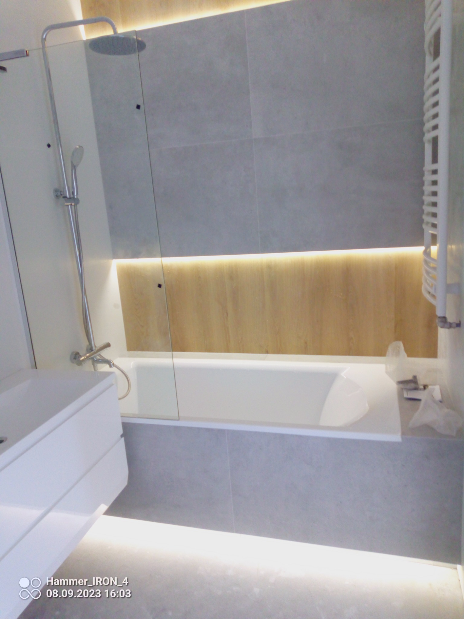 Nowoczesna łazienka z podświetlaną wanną, szarą płytką i szklanym panelem prysznicowym, widoczny biały grzejnik drabinkowy i minimalistyczna umywalka z szafką.