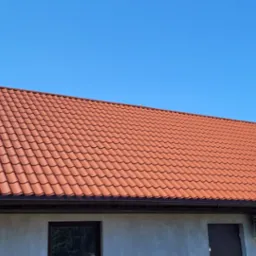 Czerwony dach z blachodachówki na domu z szarą elewacją, widoczne okna, drzwi i klimatyzator, na tle błękitnego nieba.