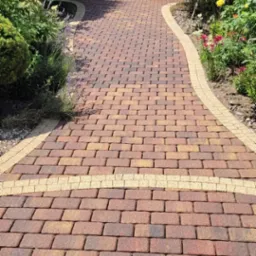 Ścieżka z kostki brukowej w odcieniach czerwieni i brązu, wykończona jaśniejszym obrzeżem, prowadząca do furtki w ogrodzeniu, otoczona bujną roślinnością z kwitnącymi krzewami.