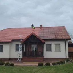 Czyszczenie dachu z czerwonej dachówki, widoczna drabina oparta o dach i pracownik w jaskrawozielonych spodniach siedzący na dachu, po prawej stronie dachu widać różnicę w czystości