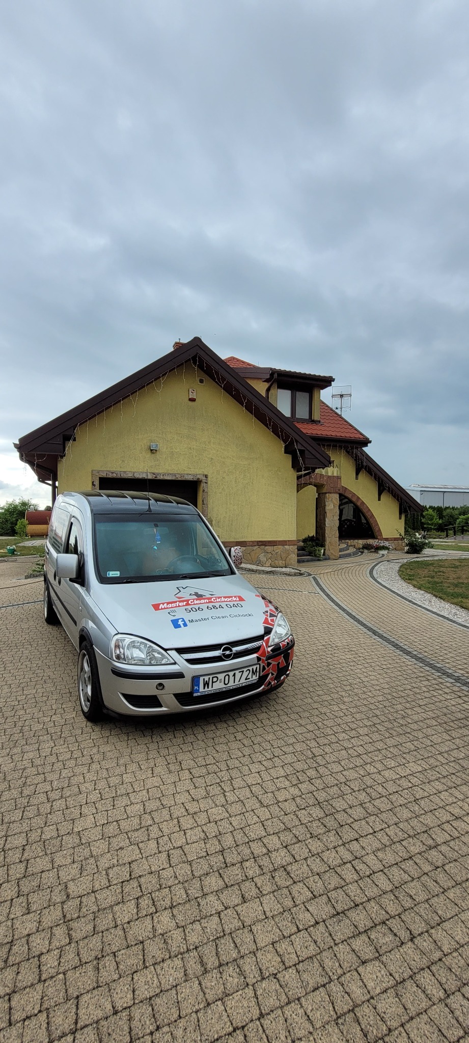 Srebrny samochód dostawczy z logo firmy sprzątającej Master Clean-Cichocki zaparkowany na kostce brukowej przed żółtym domem z brązowym dachem w pochmurny dzień.