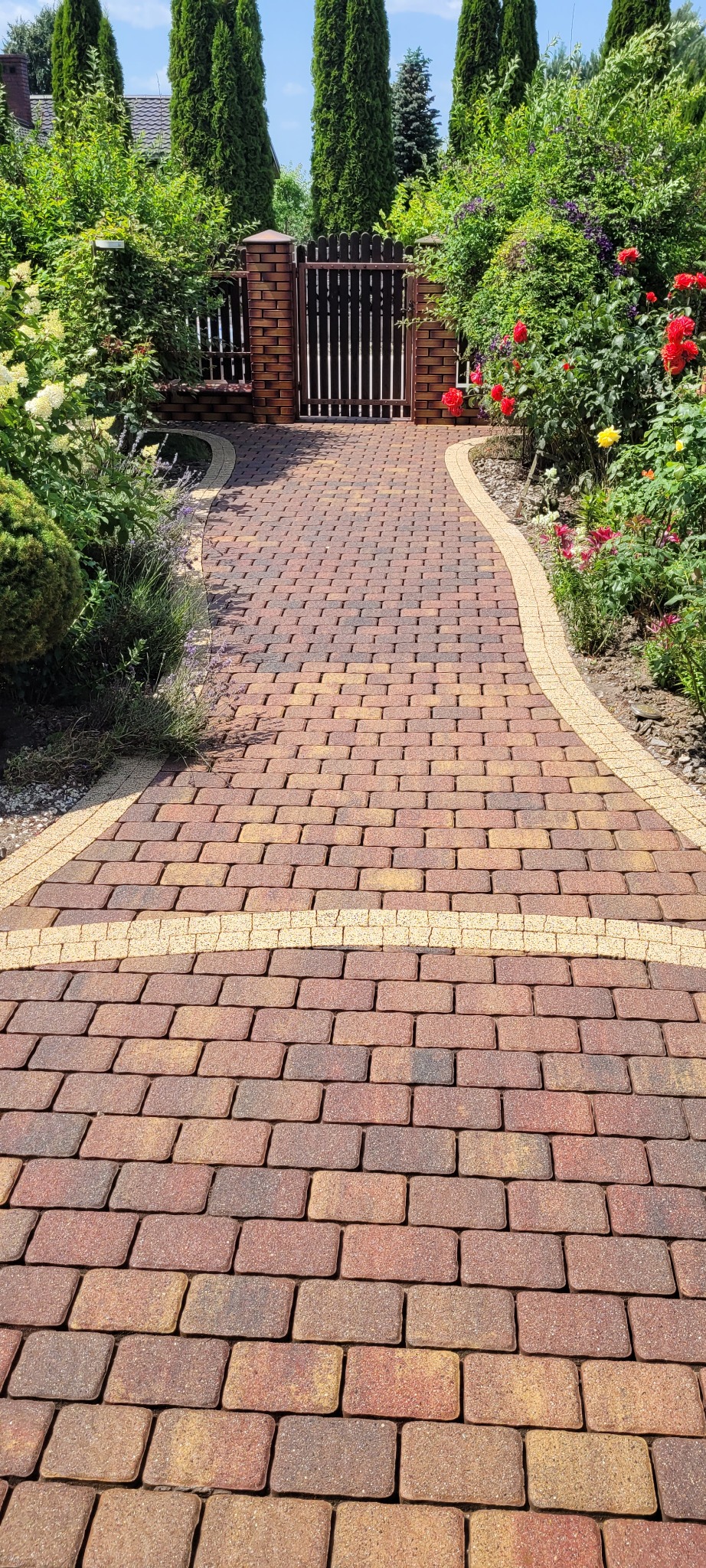 Ścieżka z kostki brukowej w odcieniach czerwieni i brązu, wykończona jaśniejszym obrzeżem, prowadząca do furtki w ogrodzeniu, otoczona bujną roślinnością z kwitnącymi krzewami.