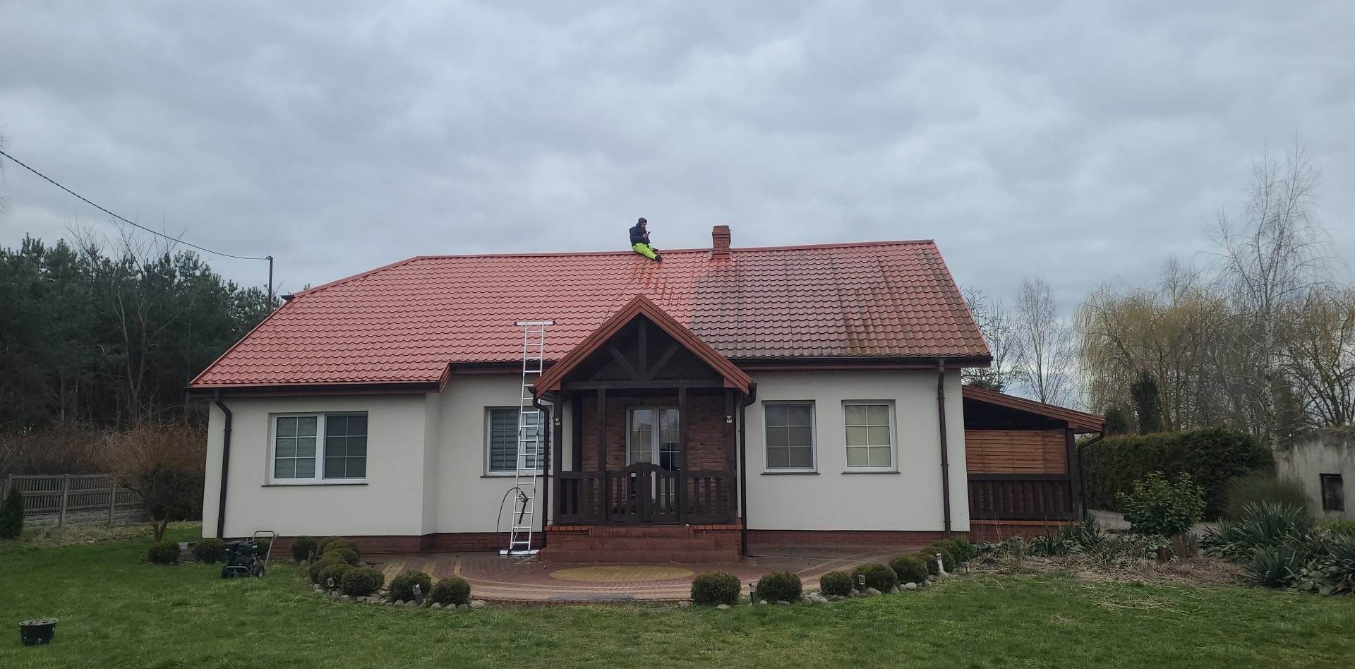 Czyszczenie dachu z czerwonej dachówki, widoczna drabina oparta o dach i pracownik w jaskrawozielonych spodniach siedzący na dachu, po prawej stronie dachu widać różnicę w czystości