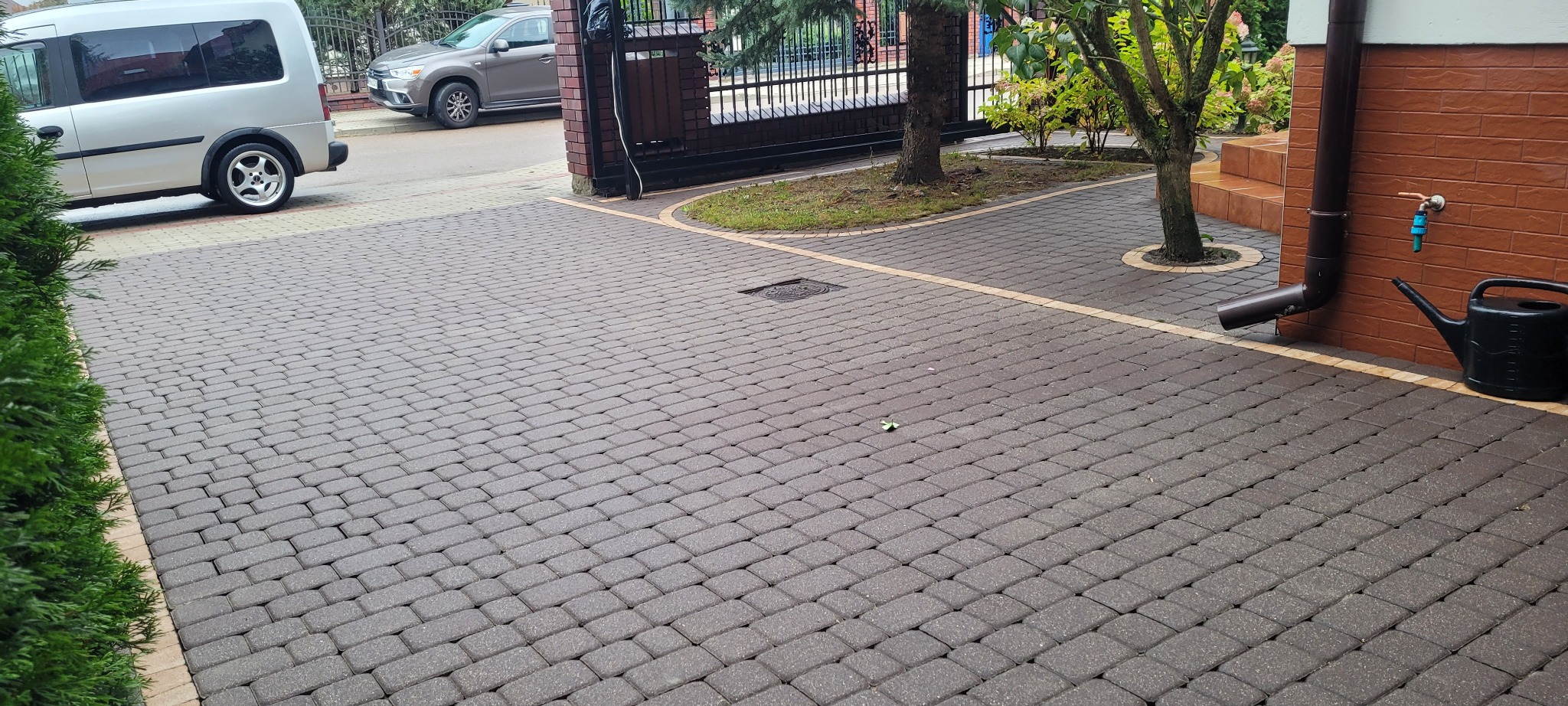 Ułożona kostka brukowa w kolorze grafitowym, z obrzeżami w kolorze piaskowym, prowadząca do bramy wjazdowej, z widoczną rynną, kranem i konewką przy ścianie budynku.