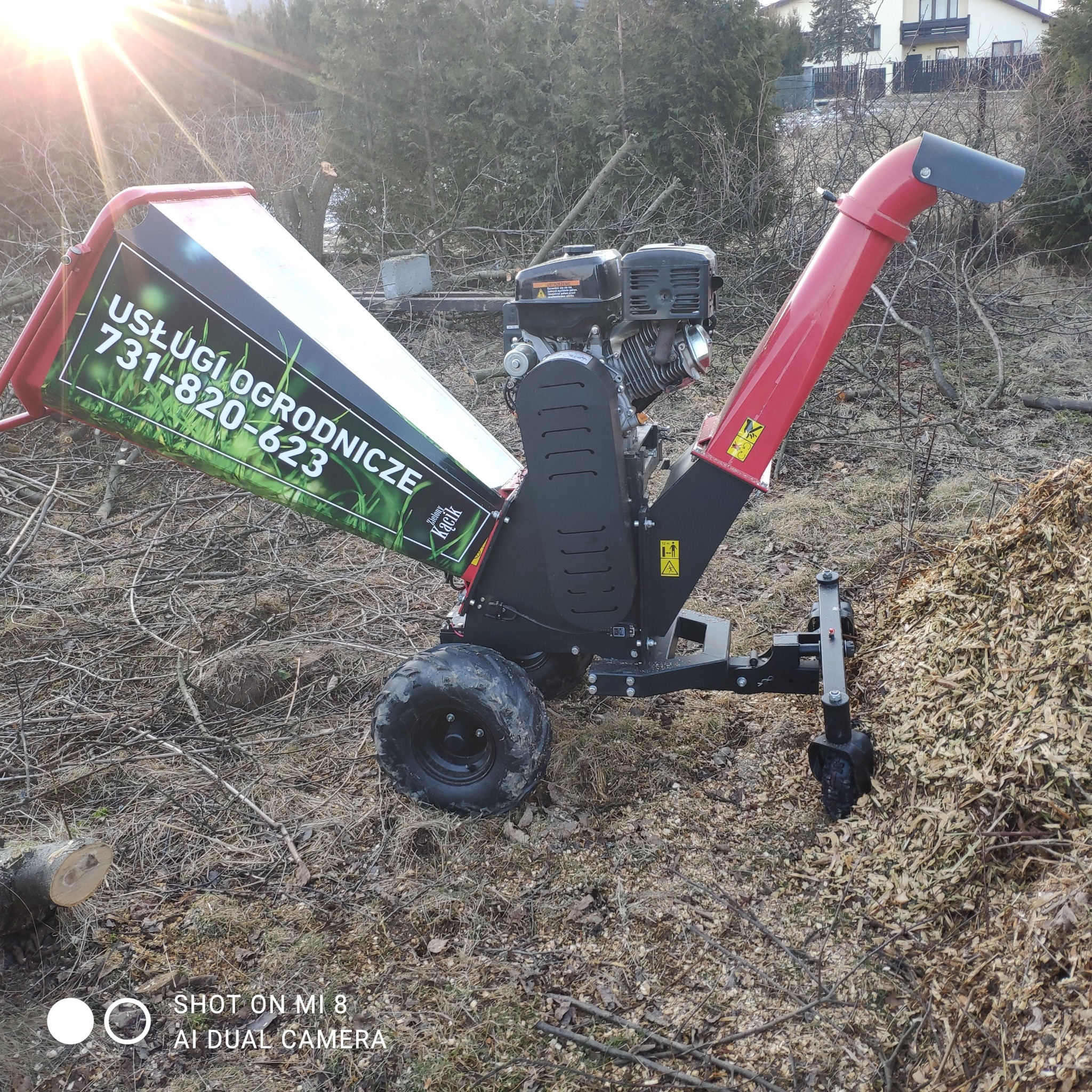 Rozdrabniacz do gałęzi z reklamą usług ogrodniczych z Bielska-Białej, na tle ściętych drzew i sterty zrębków.