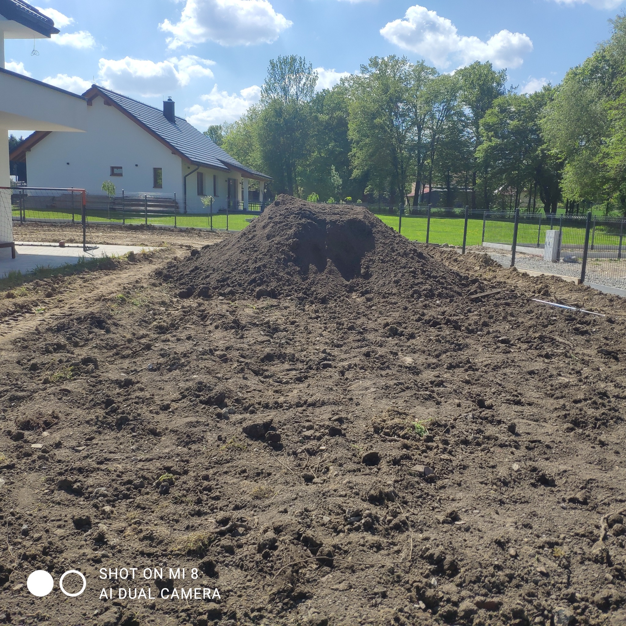Przygotowana gleba pod trawnik lub nasadzenia w ogrodzie przydomowym. Kopiec ziemi w tle, ogrodzenie panelowe i budynek mieszkalny w słoneczny dzień.