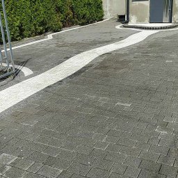 MB Przedsiębiorstwo Usługowe Maciej Mastalerczuk - Szara kostka brukowa ułożona w łukowaty wzór z jasnymi elementami, otoczona zielonymi tujami i budynkiem z szarą elewacją.