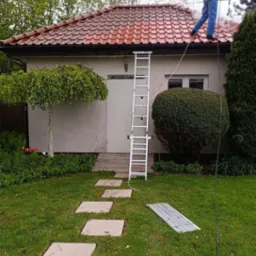 Pracownik w jeansach i kamizelce czyszczący czerwoną dachówkę myjką ciśnieniową, widoczna drabina oparta o budynek z jasną elewacją, ścieżka z płyt na trawniku.