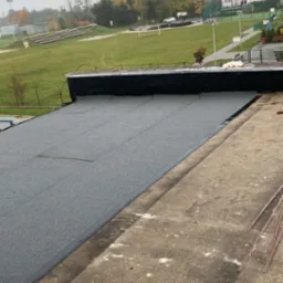 Płaski dach pokryty szarą papą termozgrzewalną, widoczne łączenia i krawędzie, w tle boisko sportowe i budynki w oddali, pochmurne niebo.